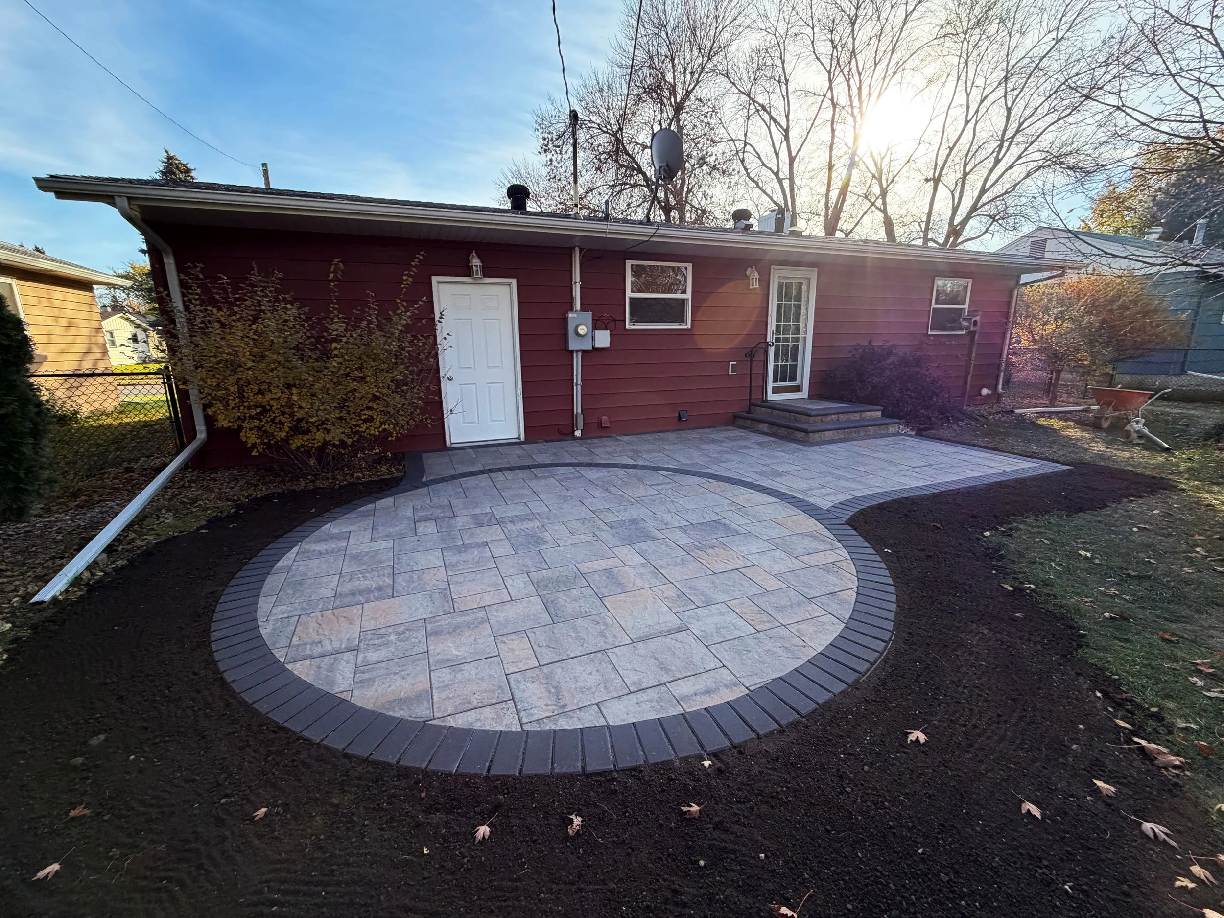 Backyard with newly installed stone patio, red house with steps and door, and a sunny sky with bare trees in fall.