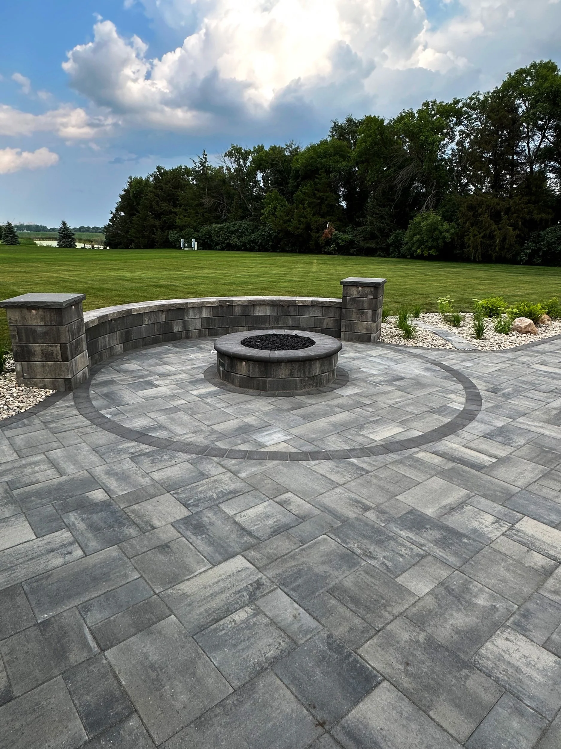 A paved outdoor patio with a fire pit in the center, surrounded by a curved stone wall and stone pillars, with a grassy lawn, trees, and cloudy sky in the background.