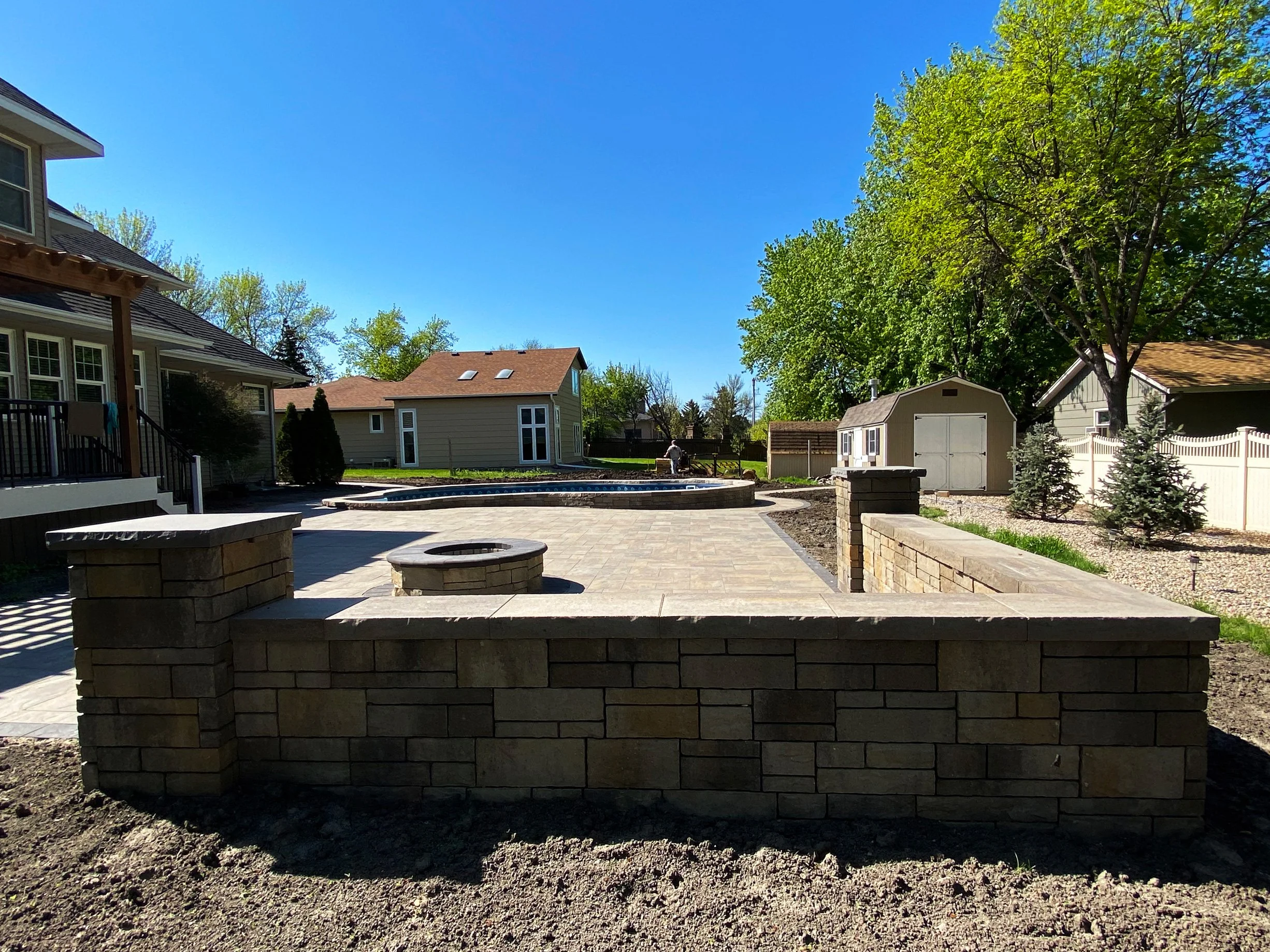 Backyard with a stone patio, fire pit, trees, shed, and neighboring houses under a clear blue sky.