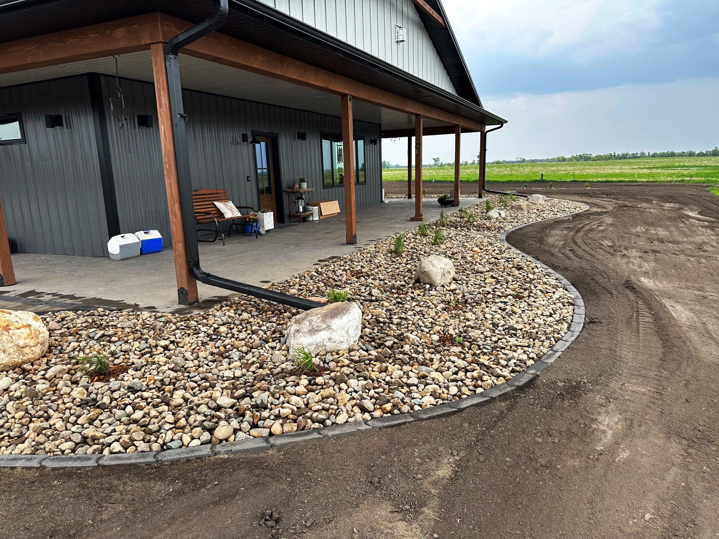 Front patio of a house with a landscaped pebble border, a bench, and some outdoor furniture, with a gravel driveway and open field in the background.