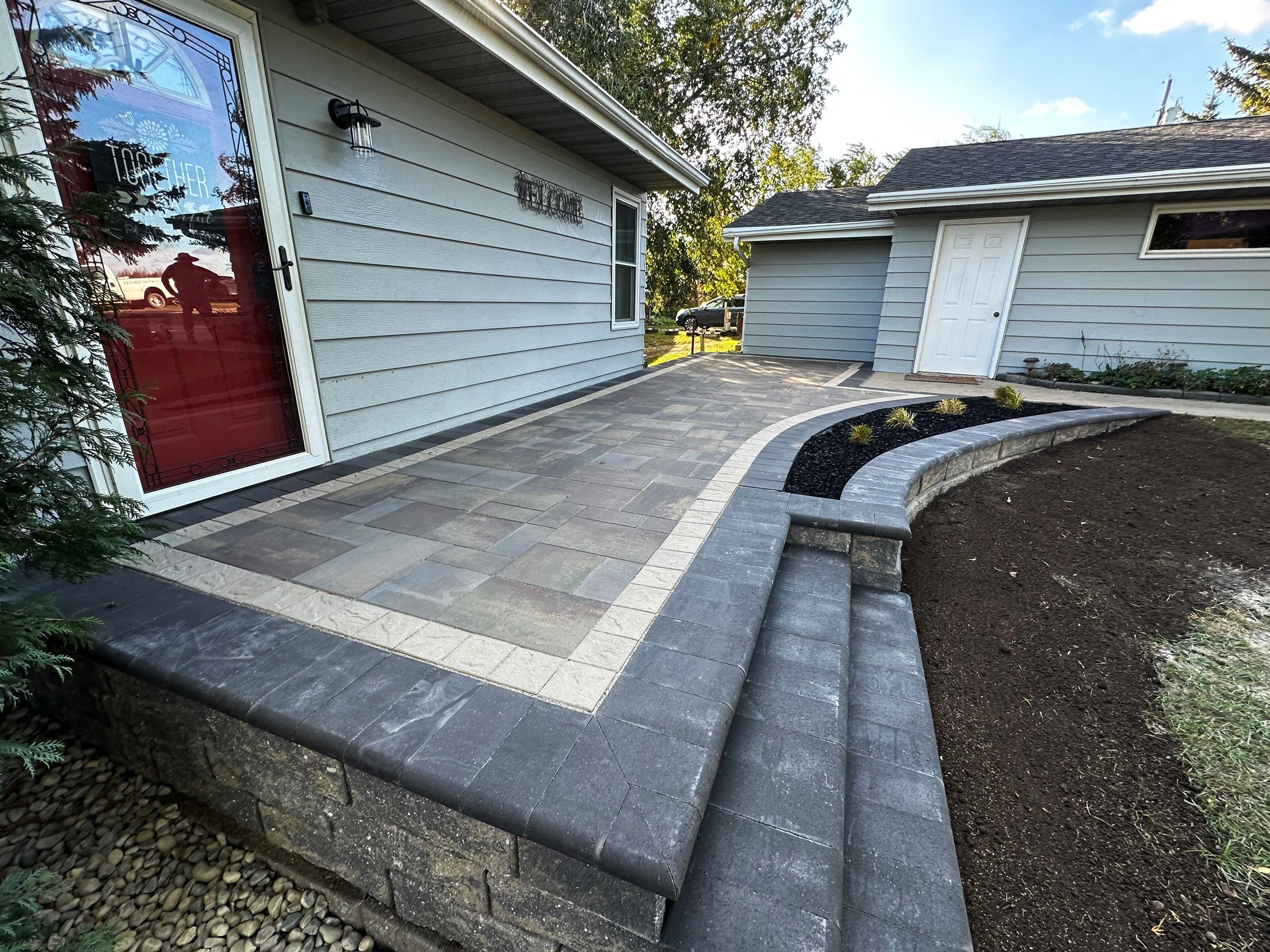 A modern backyard patio with stone pavers, curved brick edging, and decorative planting beds adjacent to a gray house.