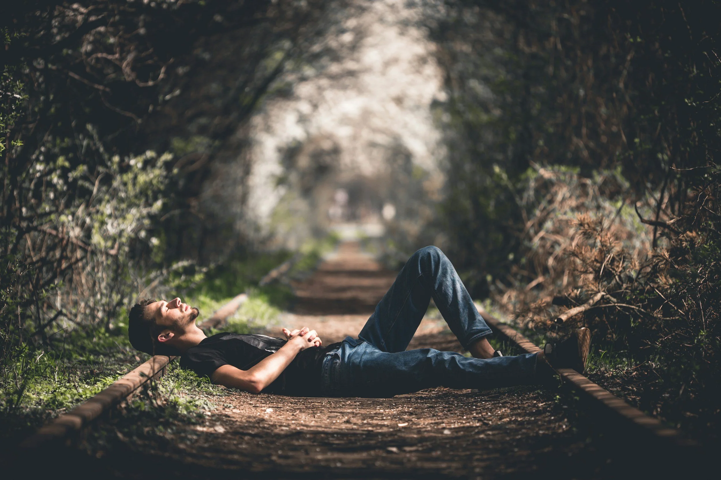A man lying on his back on abandoned train tracks in a wooded area with foliage arching overhead, eyes closed and hands resting on his chest.