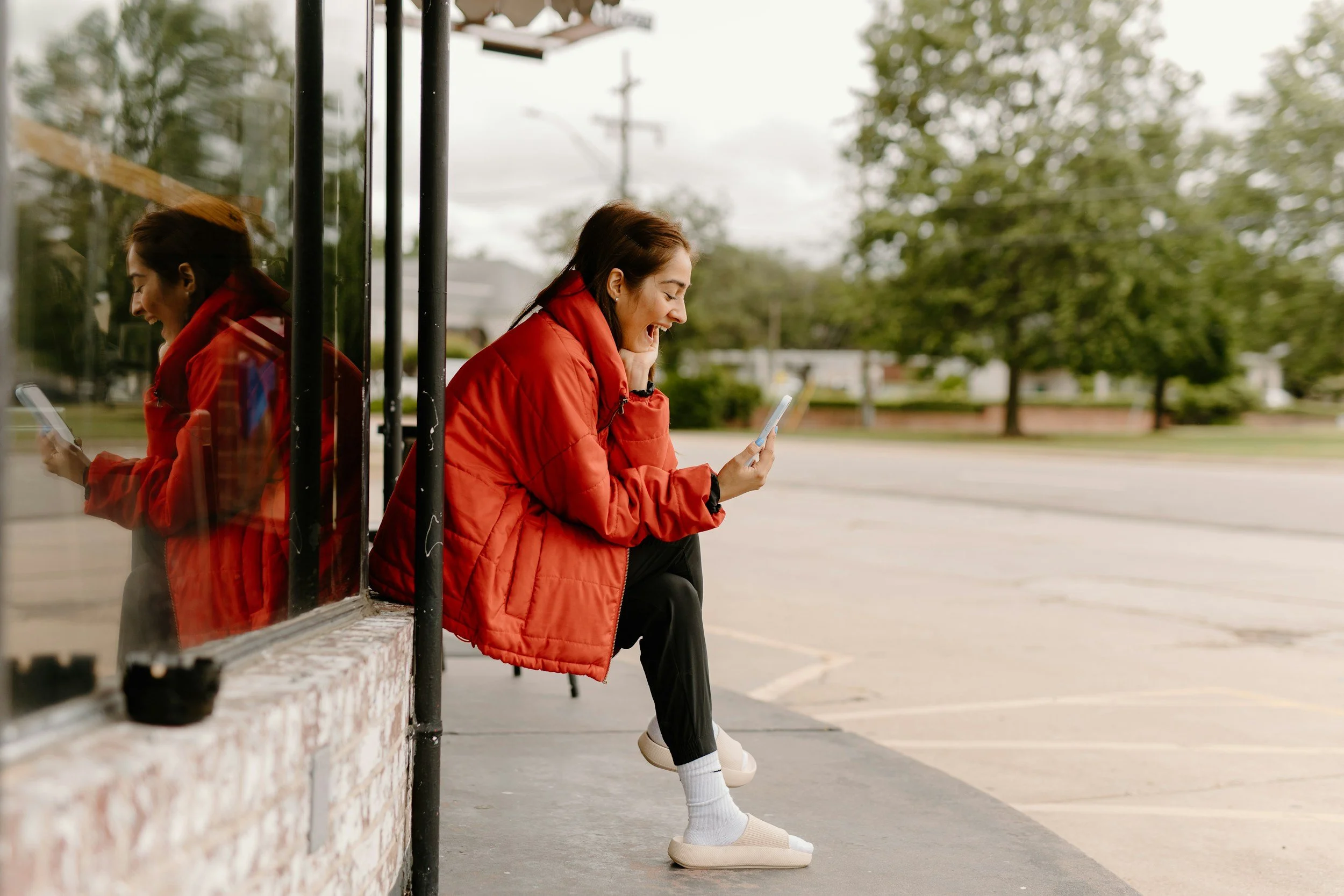 Woman in red jacket with white shoes sitting on a bench outdoors, smiling while looking at her phone, with tree-lined street in the background.