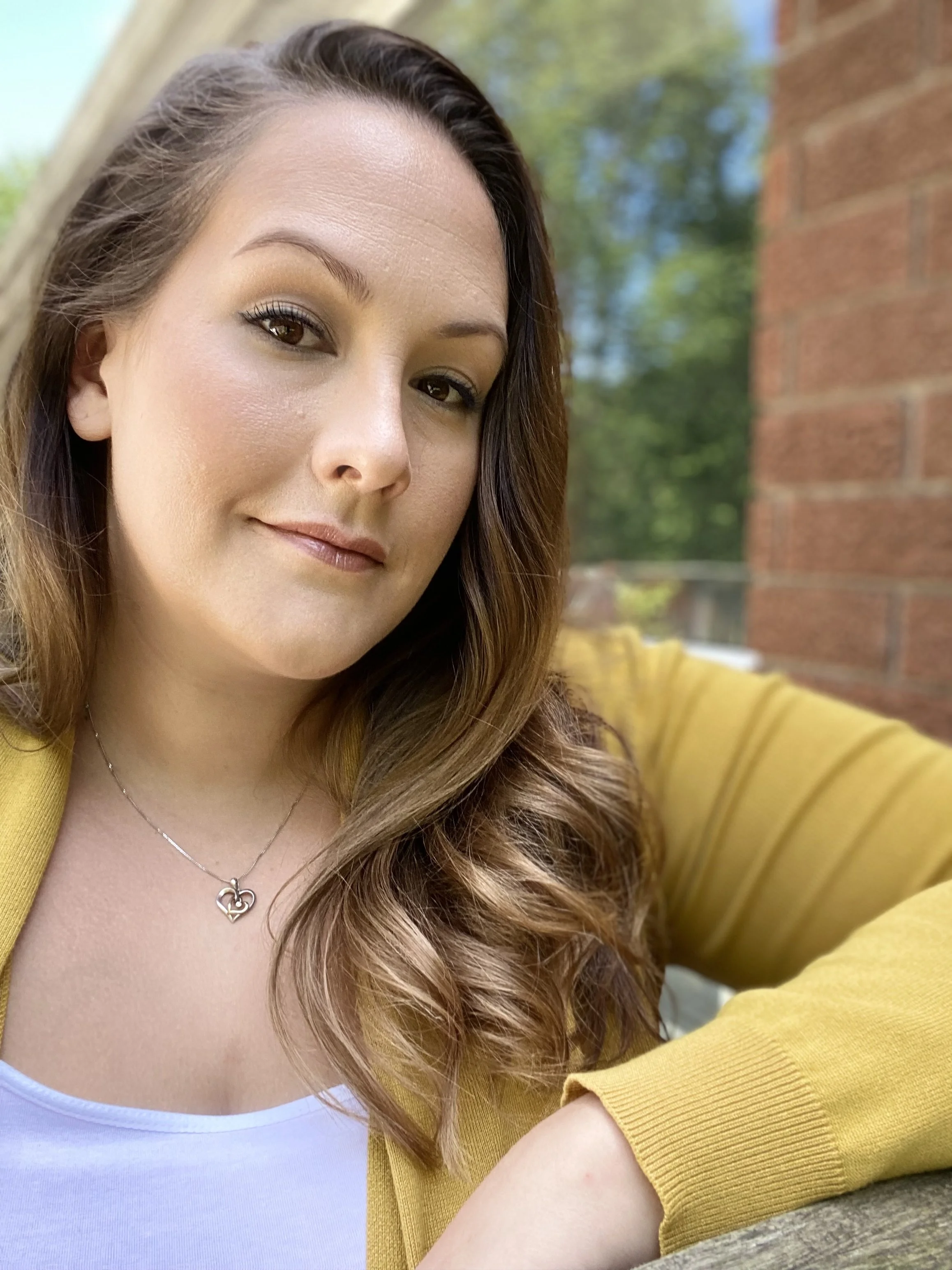 A woman with long brown hair and light makeup sitting outdoors near a brick wall, wearing a yellow cardigan, a white top, and a silver necklace with a heart-shaped pendant.