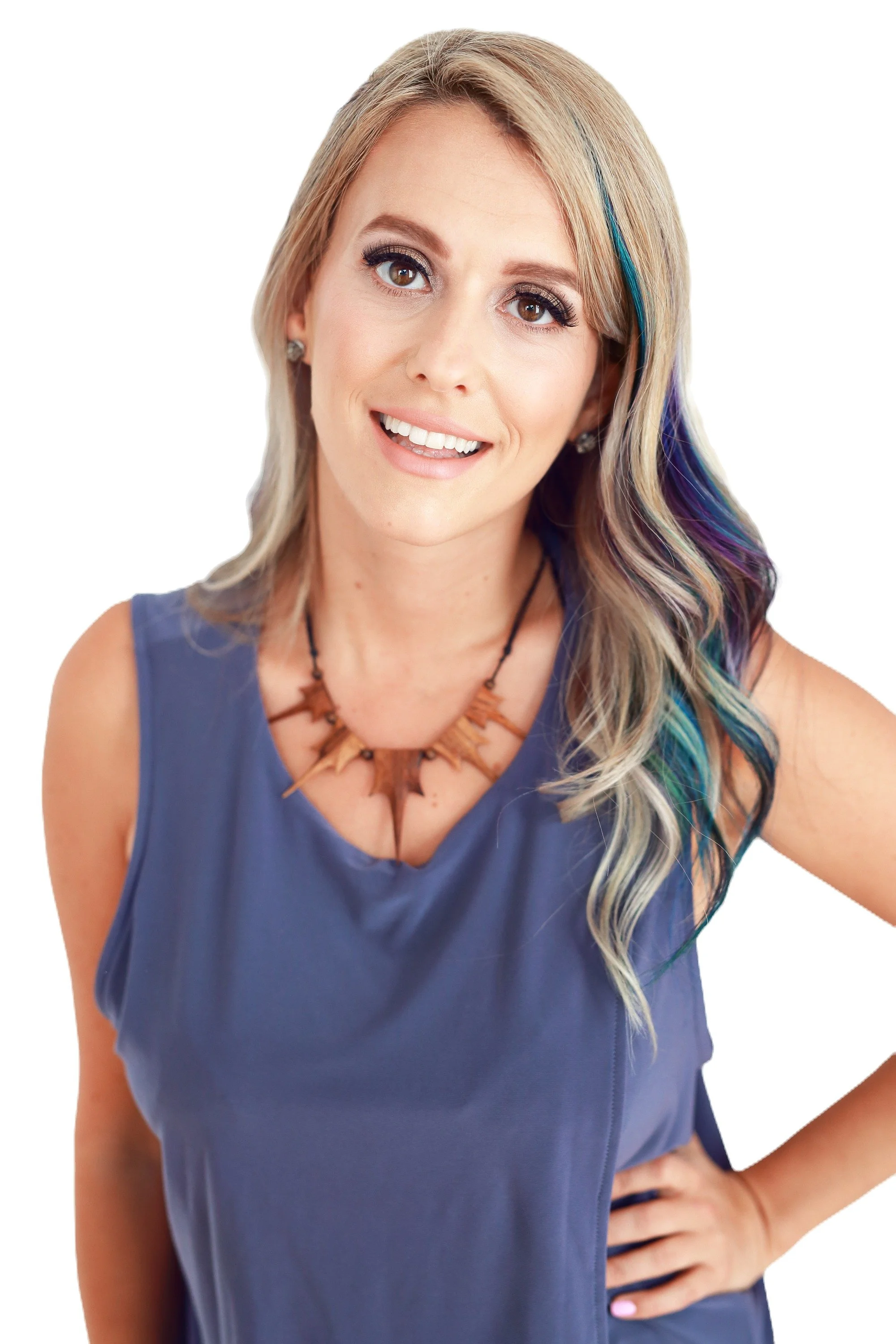 A smiling woman with long blonde hair with colorful streaks, wearing a blue sleeveless top and a wooden necklace, posing with her hand on her hip.