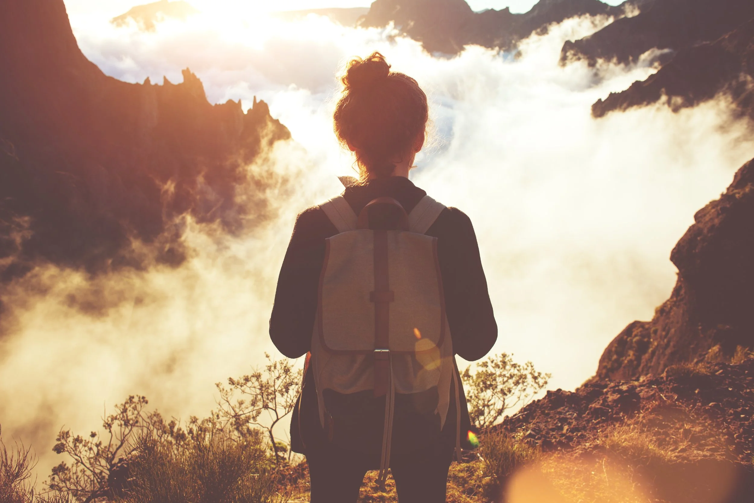 A woman with a backpack looking at a misty canyon landscape with mountains and sunrise in the background.