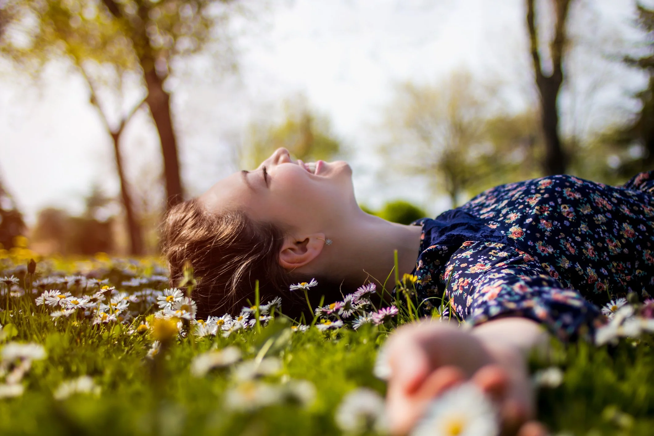A woman lying on a grassy field filled with small white flowers, smiling and enjoying the sunshine with trees in the background.