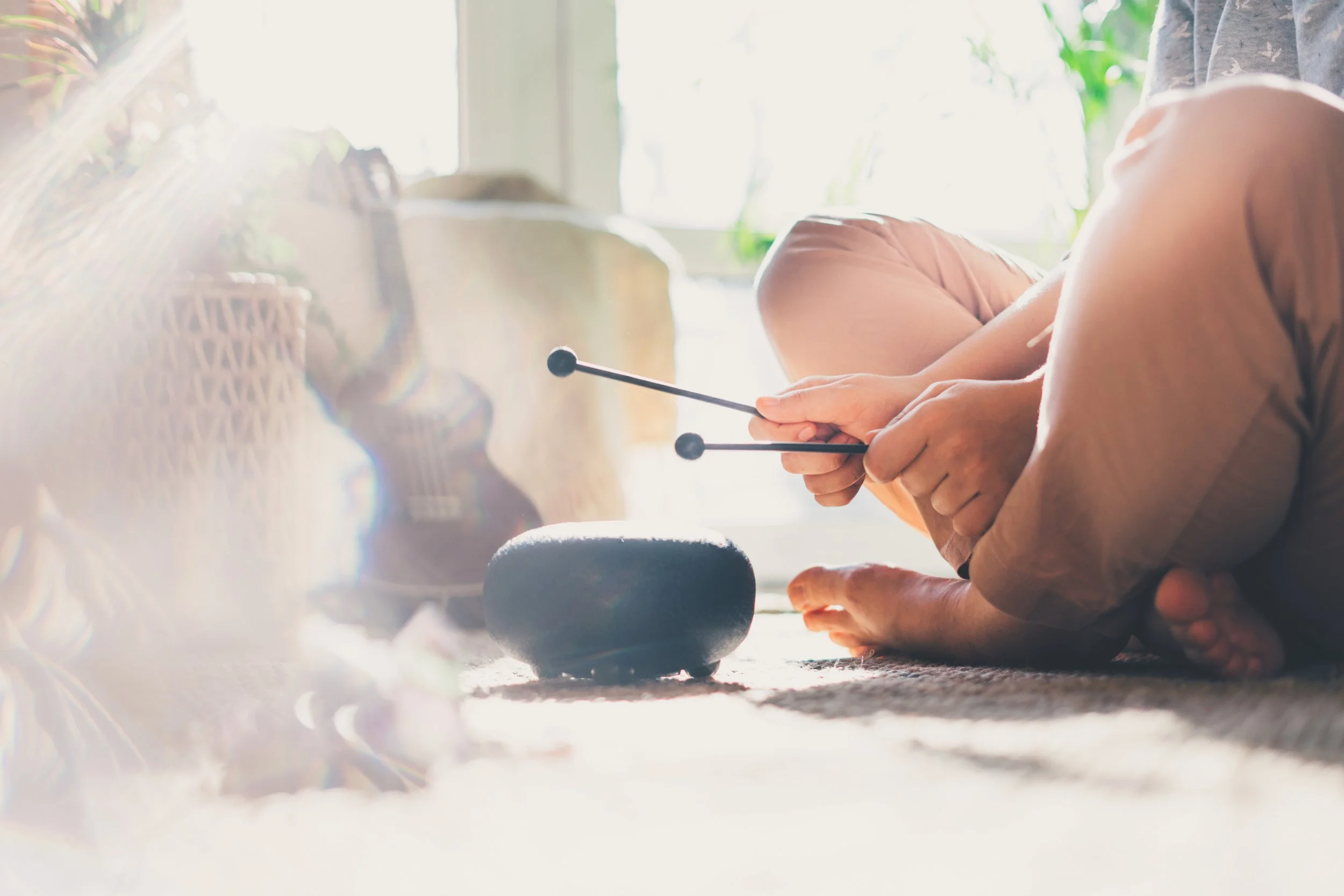 Person lying on the floor, holding tuning forks near a singing bowl, indoors near a window with sunlight, plants, and a guitar in the background.
