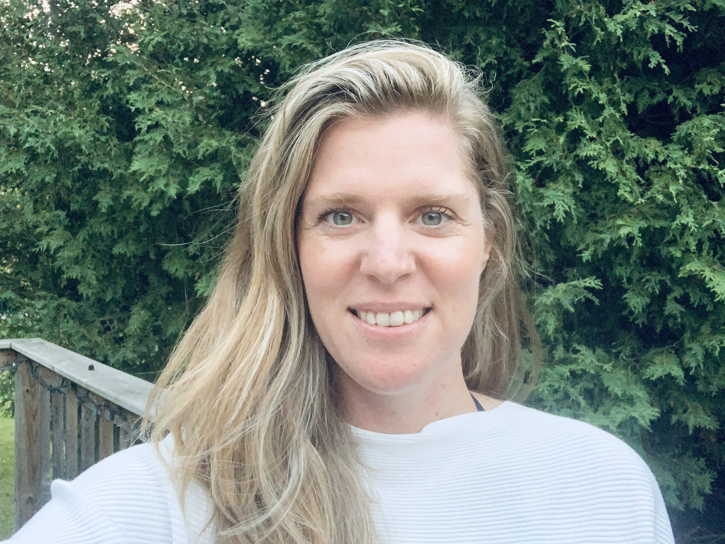 A woman with long blond hair, smiling, wearing a white top, outdoors with green trees in the background.
