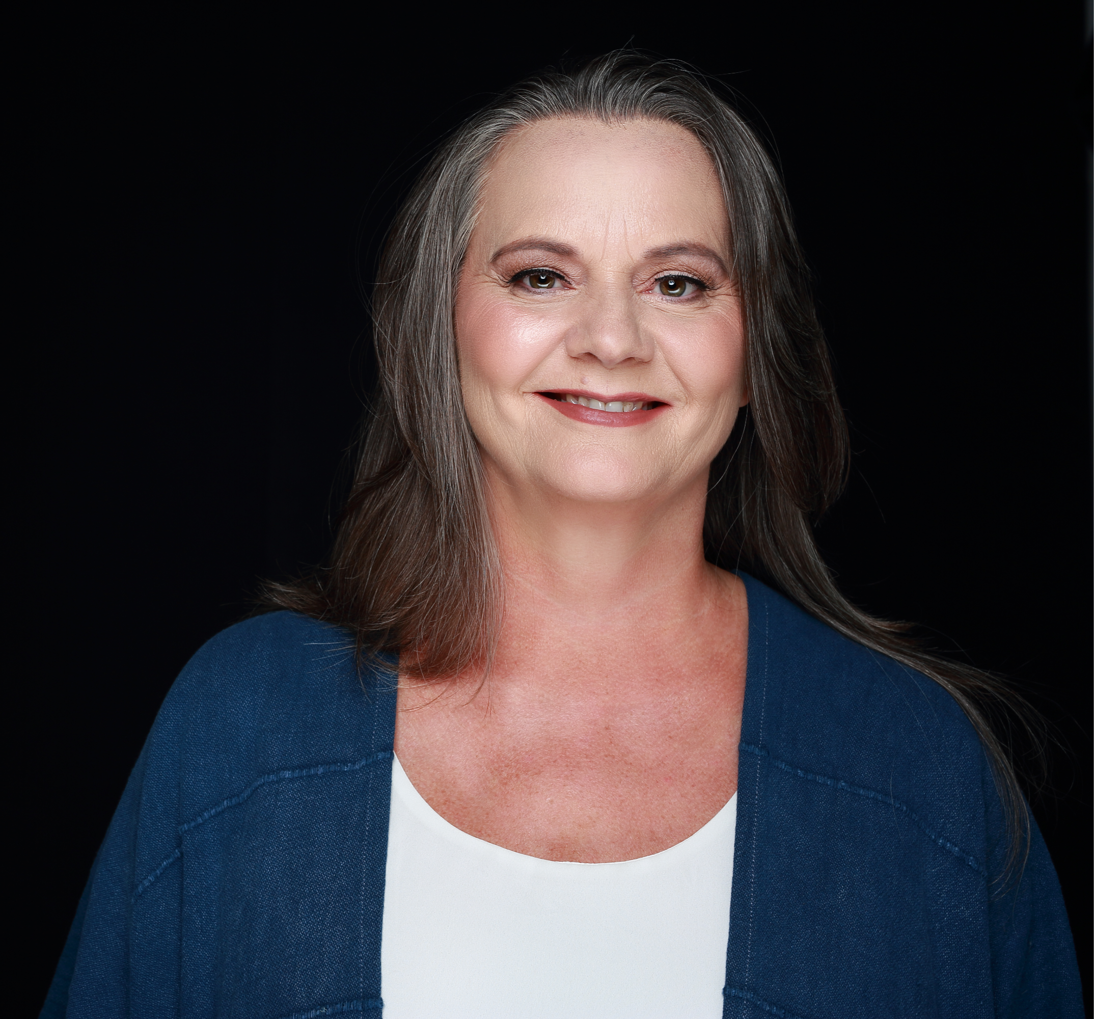 A smiling middle-aged woman with long gray hair, wearing a white shirt and a blue jacket, against a black background.