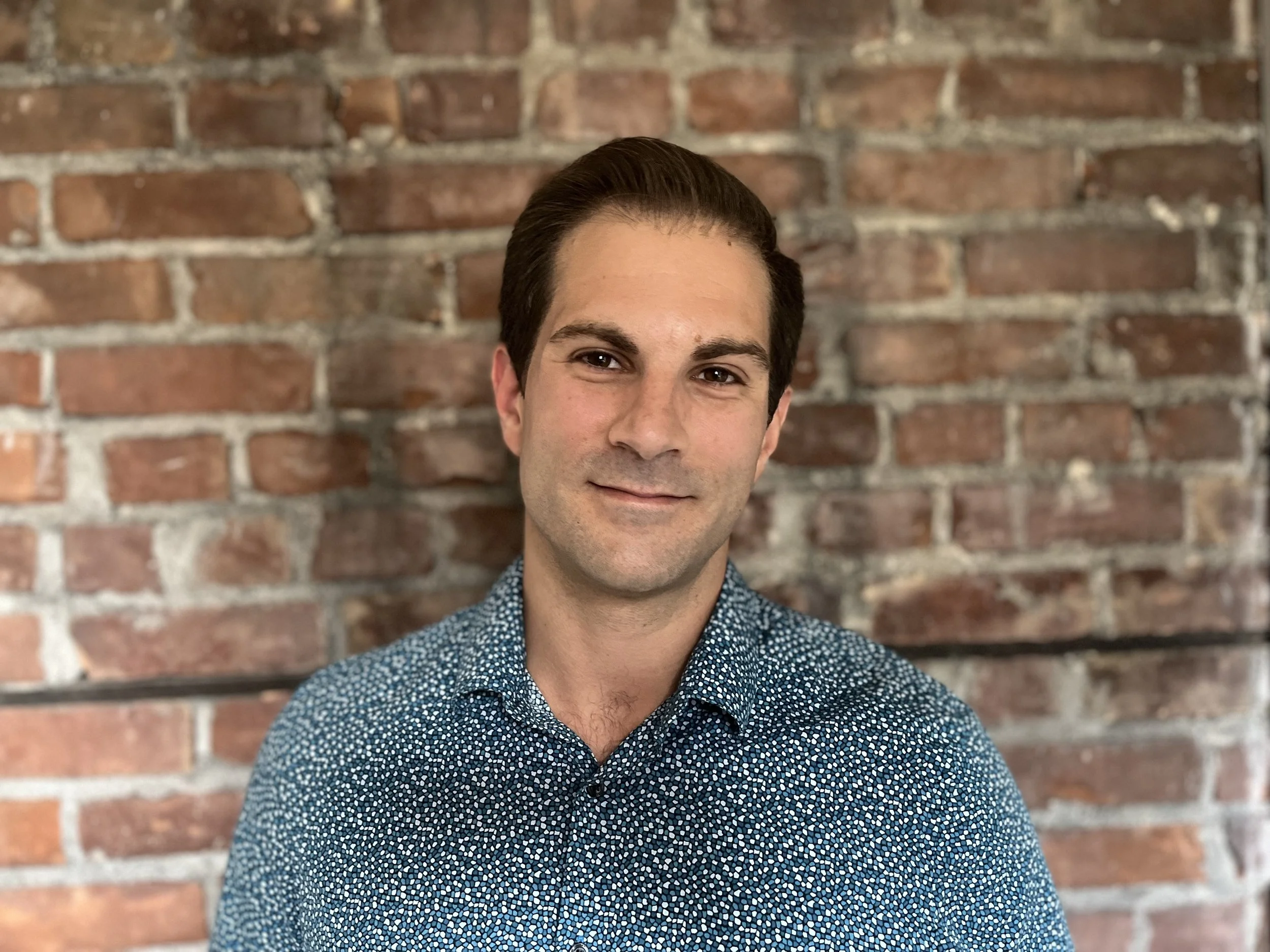 A young man with dark hair styled back, wearing a blue patterned button-up shirt, standing in front of a brick wall.