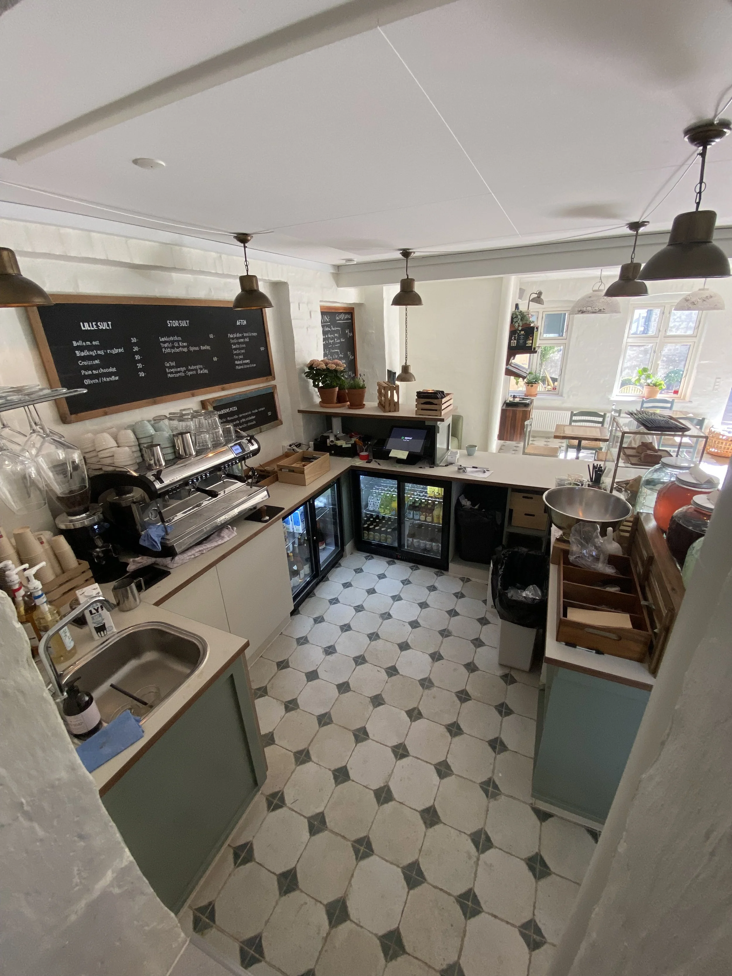 Inden for en café eller café-lignende sted med sort tavle med menupunkter, kaffemaskine, køleskab med drikkevarer, små borde og vinduer med planter.