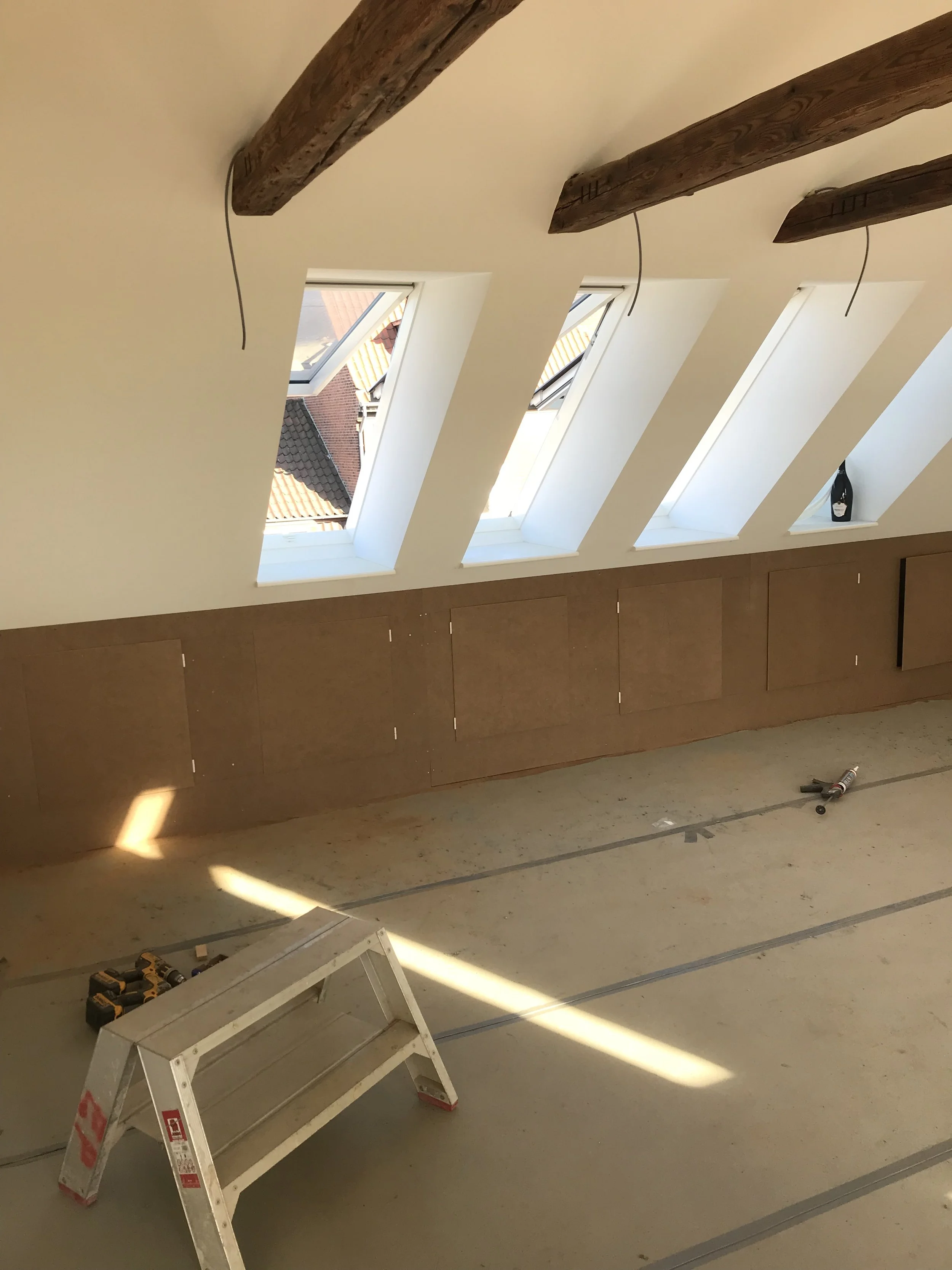 Værelse under renovering med tre ovenlysvinduer, træbjælker i loftet, værktøj på gulvet og en skumkanon på gulvet.