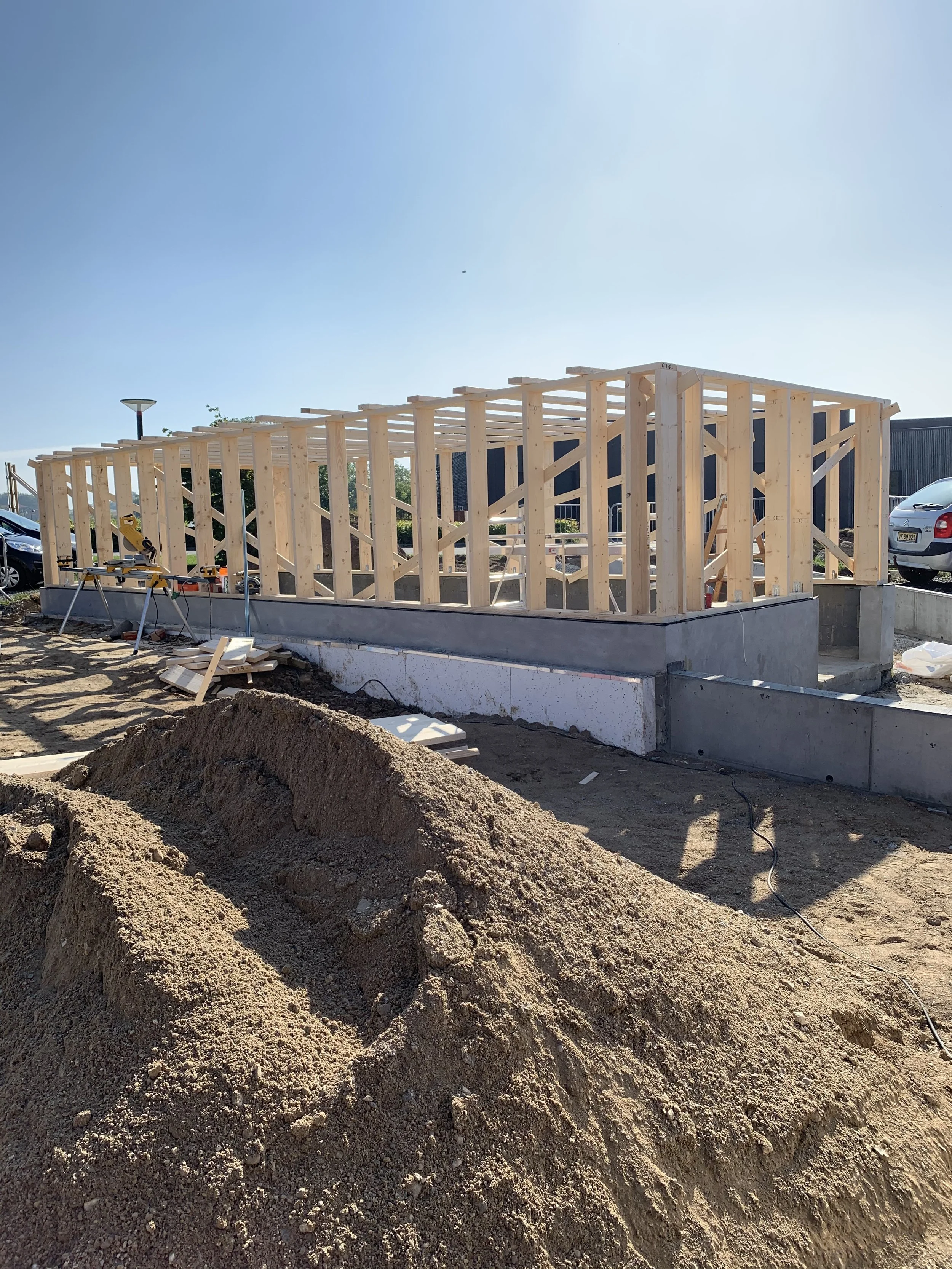 Et under opførelse konstruktion med trækonstruktion, bygget på en betonfundament. Der er bygget på en byggeplads med jord og materialer rundt om.