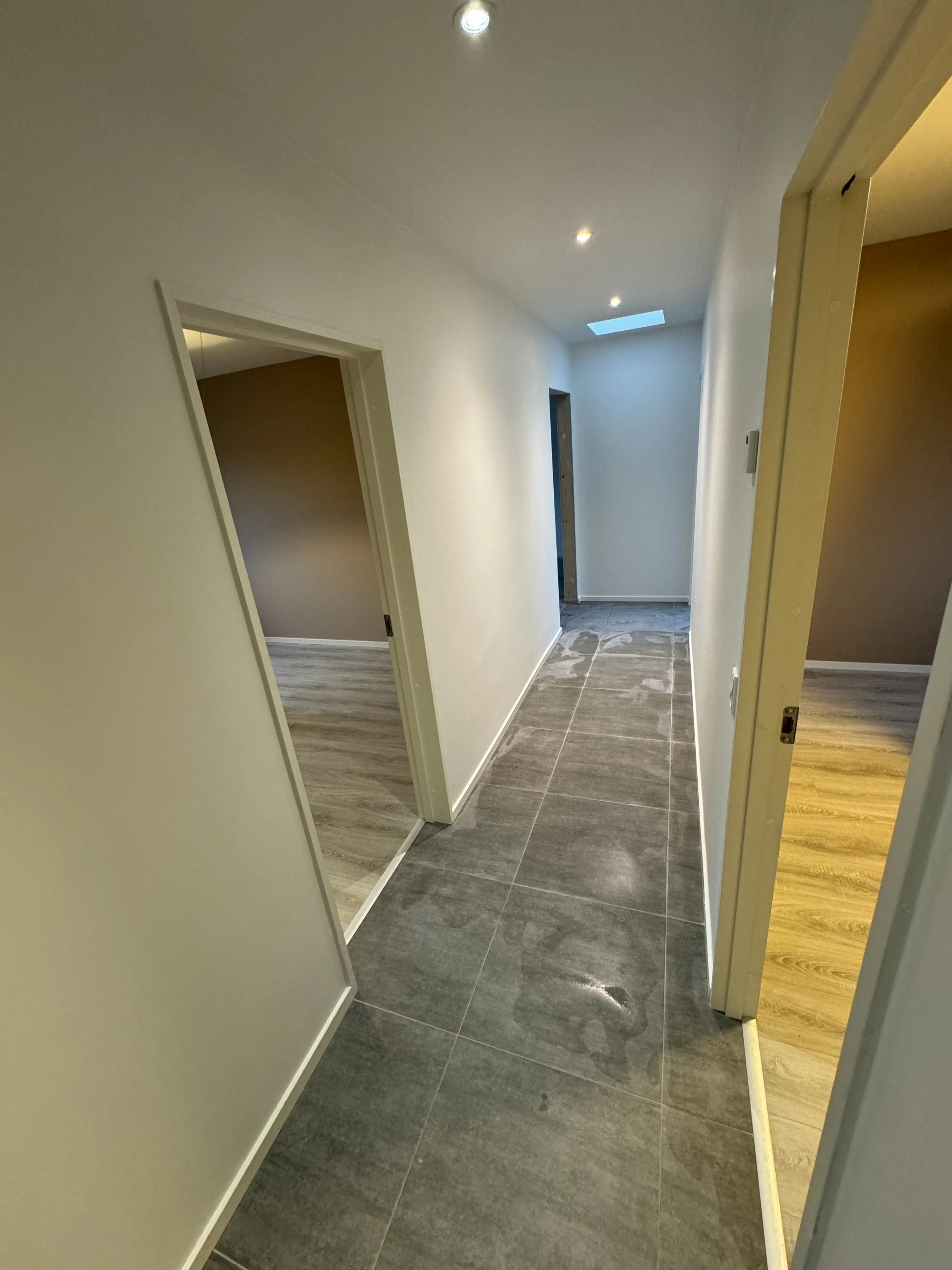 A hallway with three doorways, gray tile floor, off-white walls, recessed ceiling lights, and a skylight.