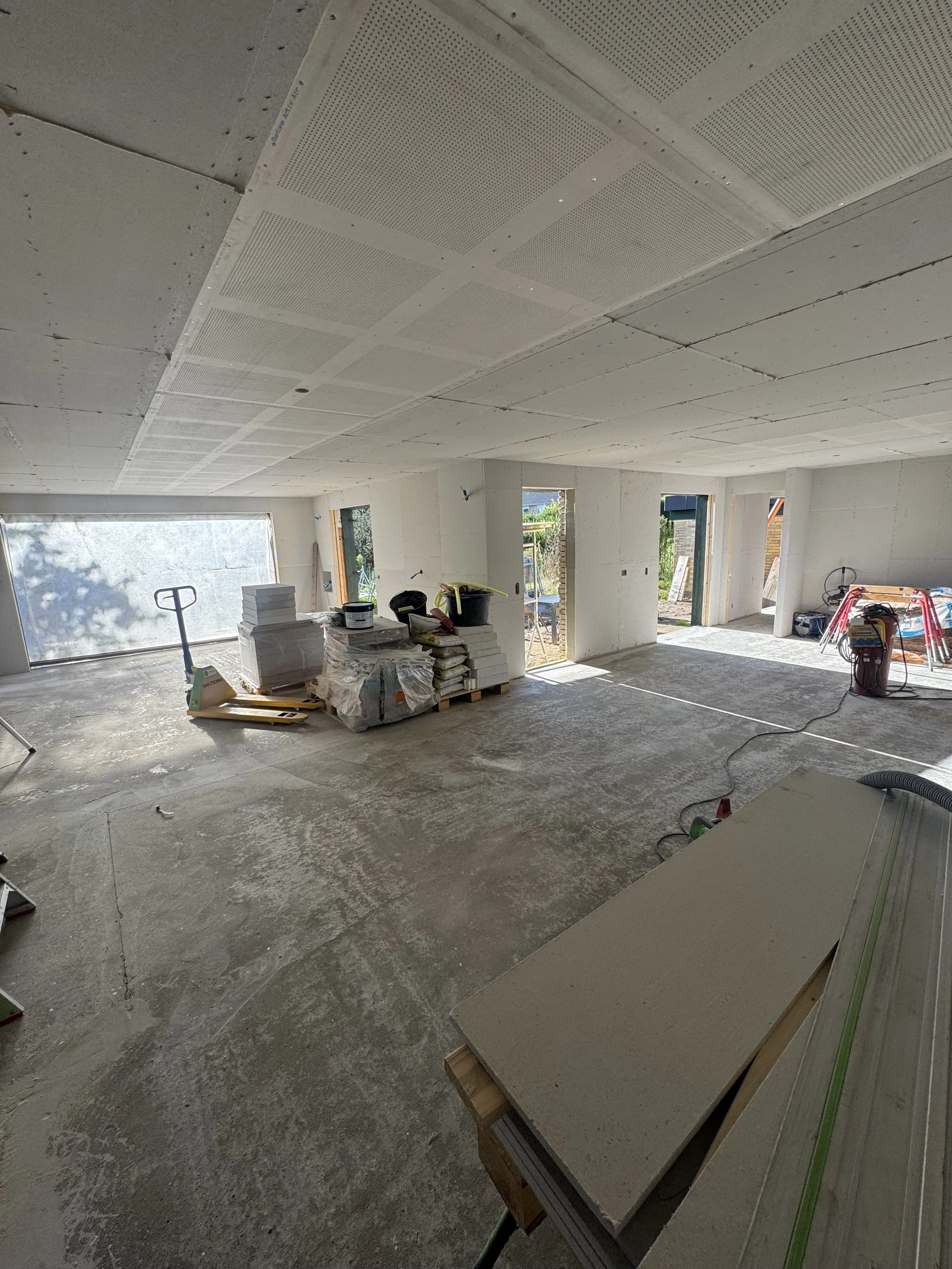 Indendørs byggeri med ikke færdighærdig betonboden, materialer og værktøj spredt rundt, store vinduer og åbne døre, hvilket antyder at konstruktionsarbejde stadig er i gang.