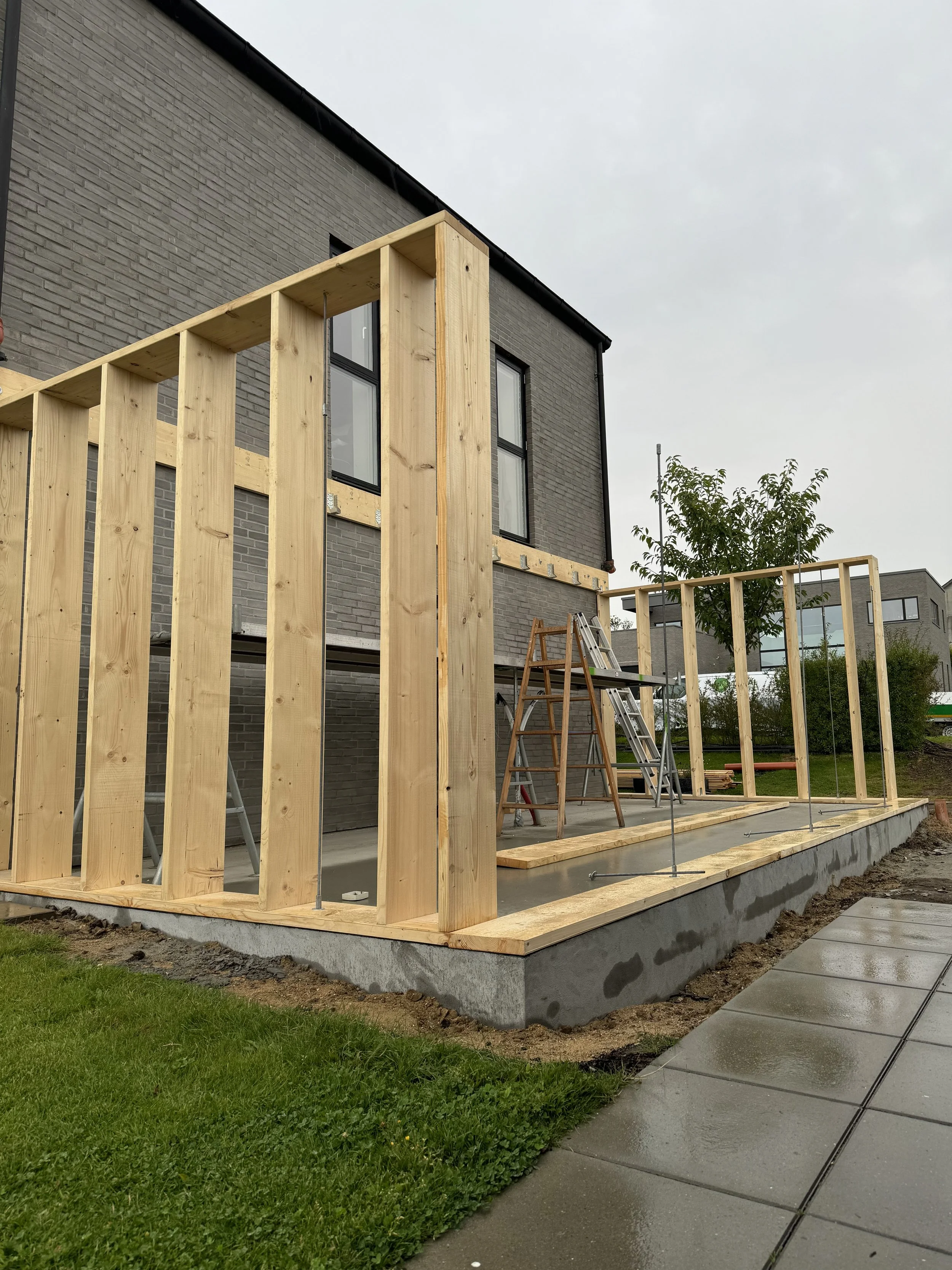 En bygning under konstruktion med træbindingsværk og en murstensfacade. Der er stilladser, en stige og lærred på området, og den omgivende have er grøn med en sti.