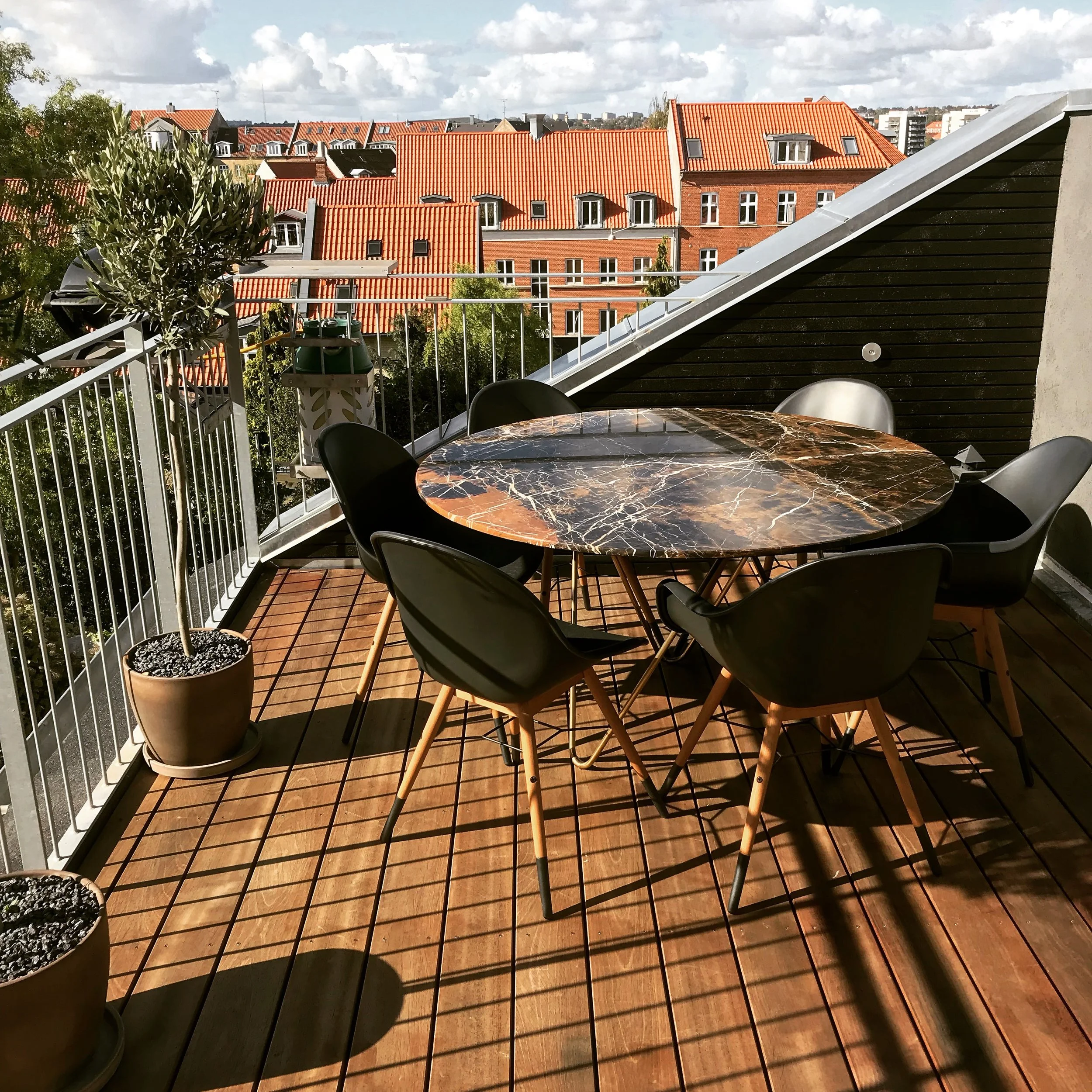 Udefra et balkon med et rundt bord og fire stole, planter og rister til tøj med en udsigt over røde tegltage og bygninger under en skyet himmel.