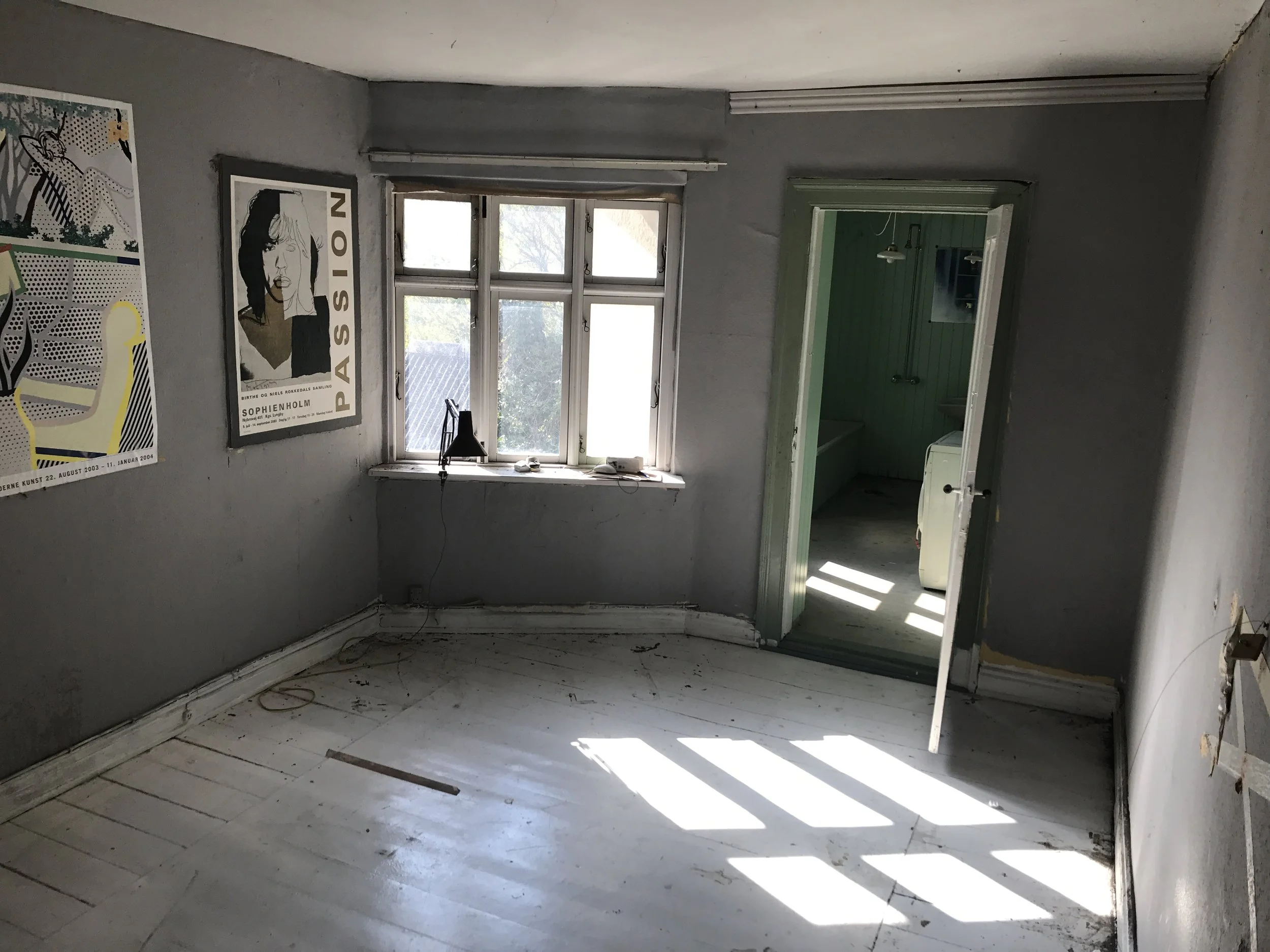 Et tomt og forladt værelse med grå vægge, en stor vindue, der lukker lys ind, og en åben dør, der fører til et andet rum med grøn væg. Der hænger to plakater på væggen, en af Sophie Hall og en anden med en tegning af en kvinde.