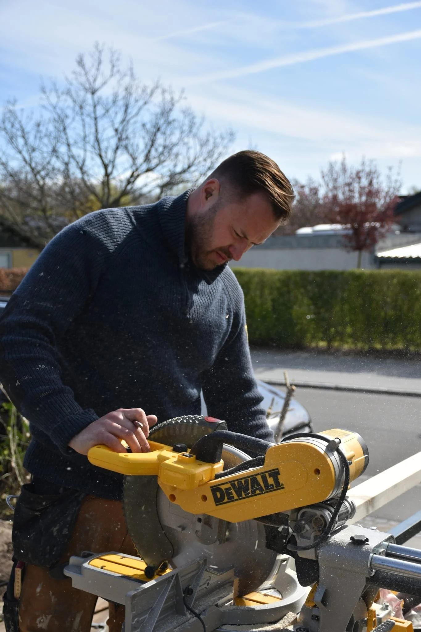 Mand med kort, brunt hår klipper en plade med en DeWalt rundsav udendørs på en solskinsdag