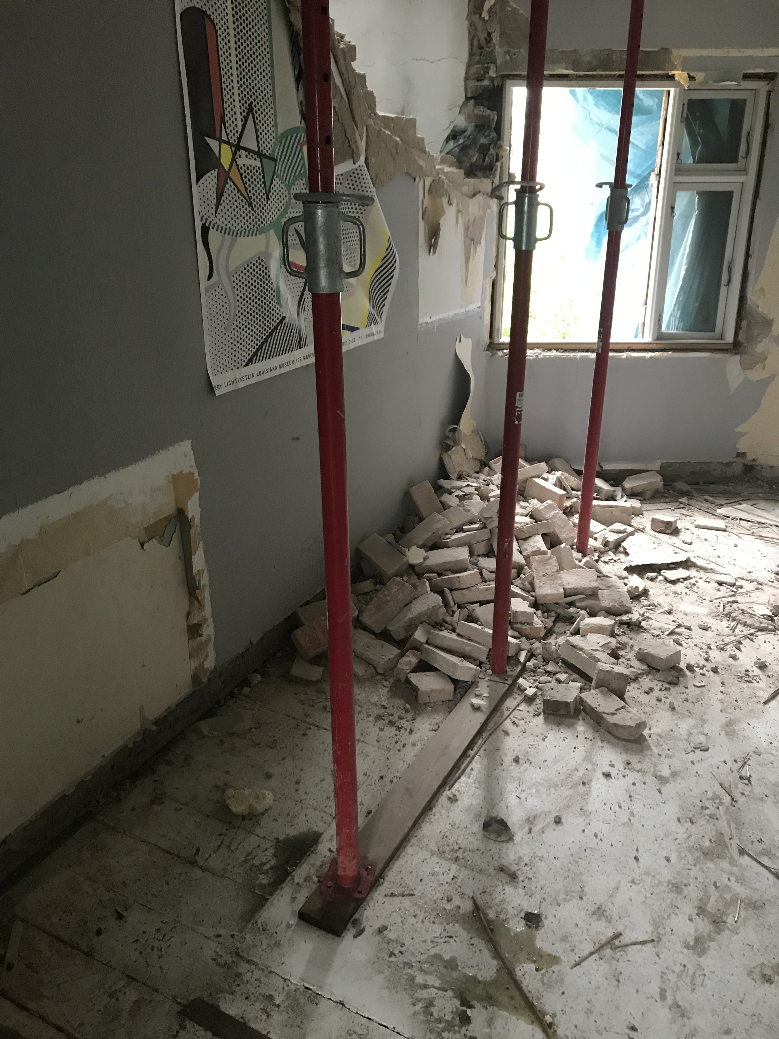 Et rum under renovering med nedbrudte vægge, murbrokker på gulvet, og røde støtteelementer til byggearbejde.