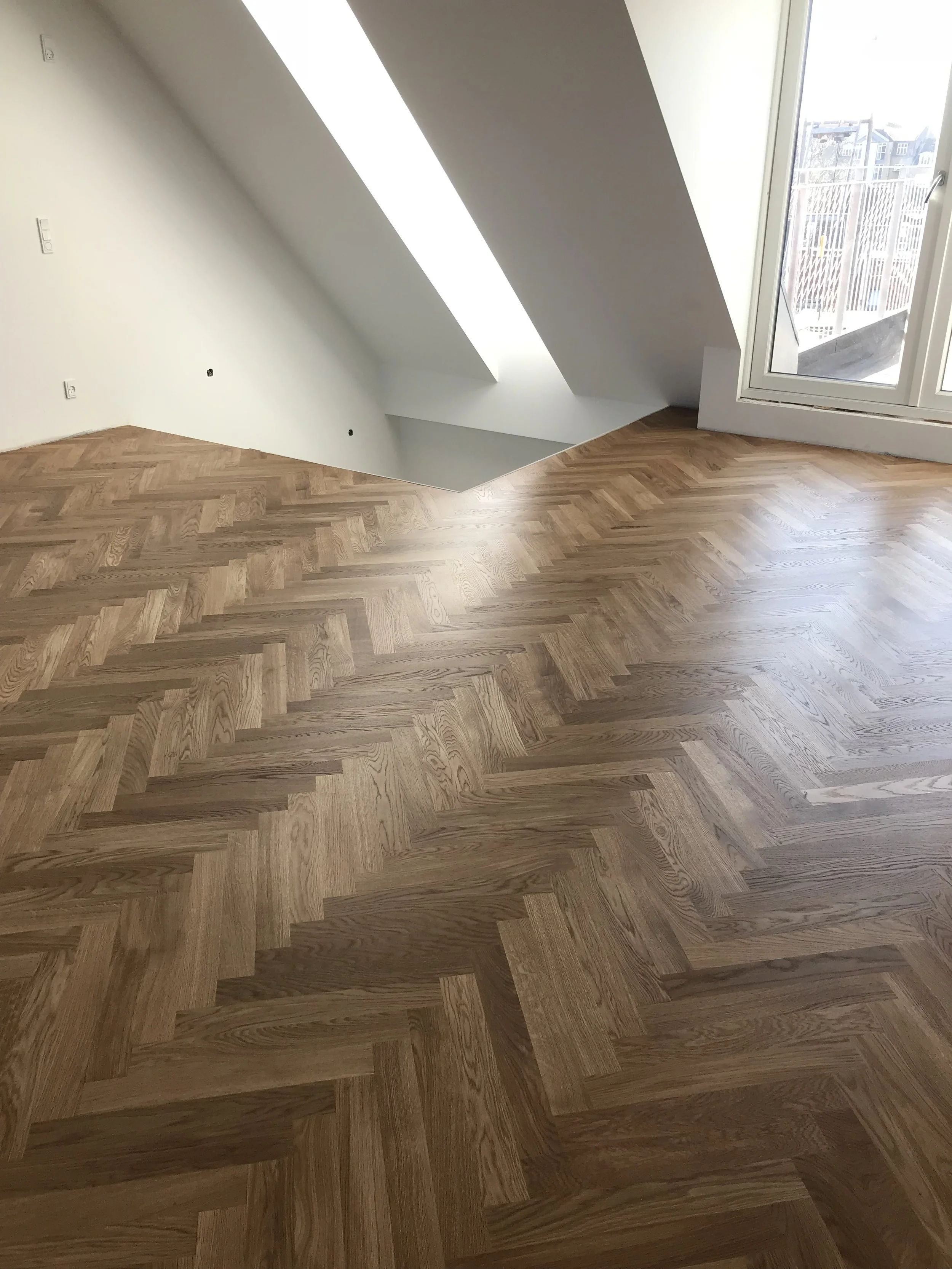 Indendørs billede af et rum med trægulv, en stor vindue på højre side og en væg med et hul, der sandsynligvis er under renovering.