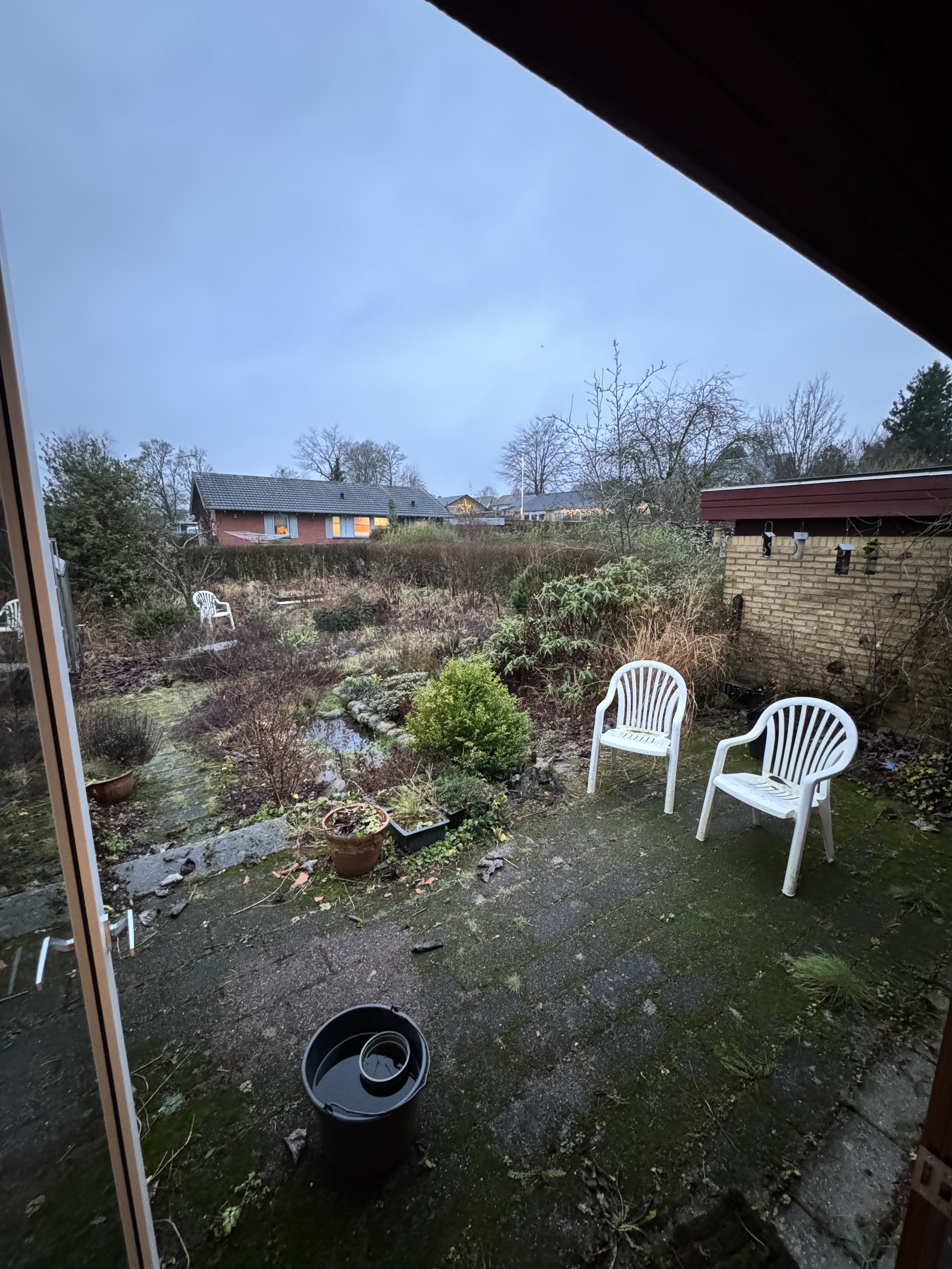 Udsigt fra et hus, hvor en have er lidt rodet med potter, græs, buske, og tre hvide plaststole. Der er også en sort affaldskurv, og udsigten viser andre huse med tagsten og træer i baggrunden mod en klar blå himmel.