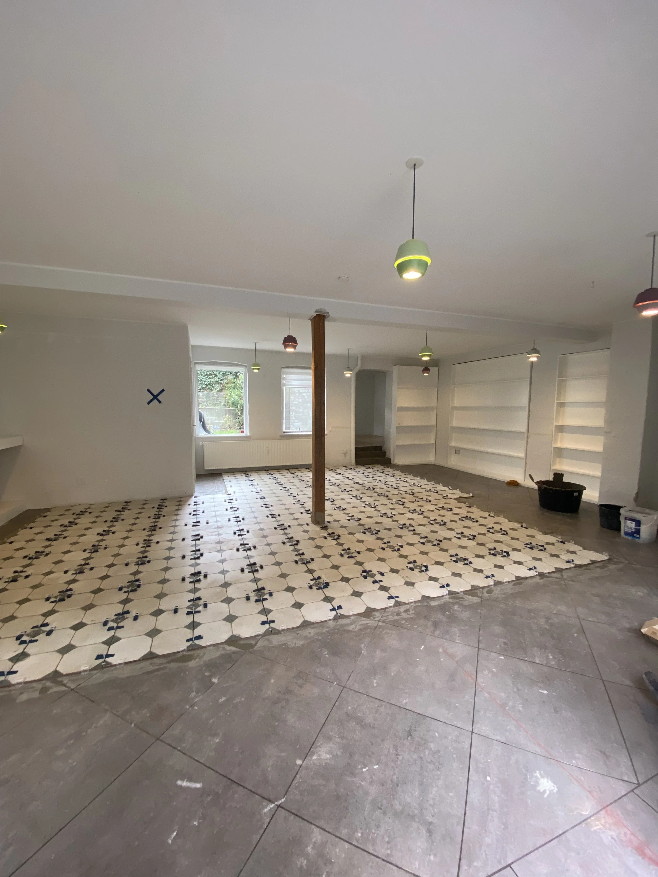 Indendørs rum under renovering med nye fliser, der er ved at blive lagt, og ophængte lamper i loftet.