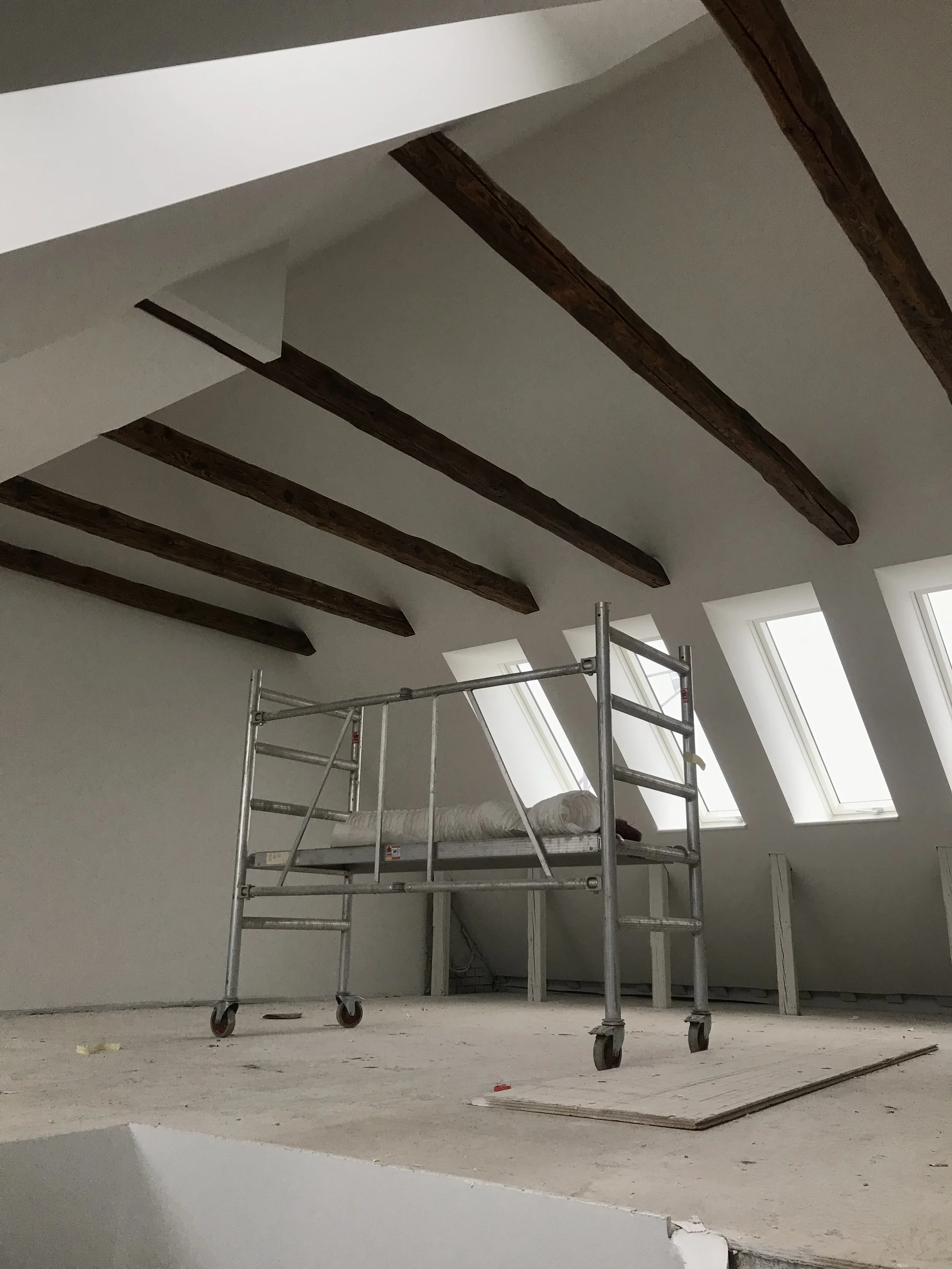 Indendørsunder renovation med en midlertidig arbejdsplatform og træbjælker i loftet samt tre vinduer, der lader naturligt lys ind.