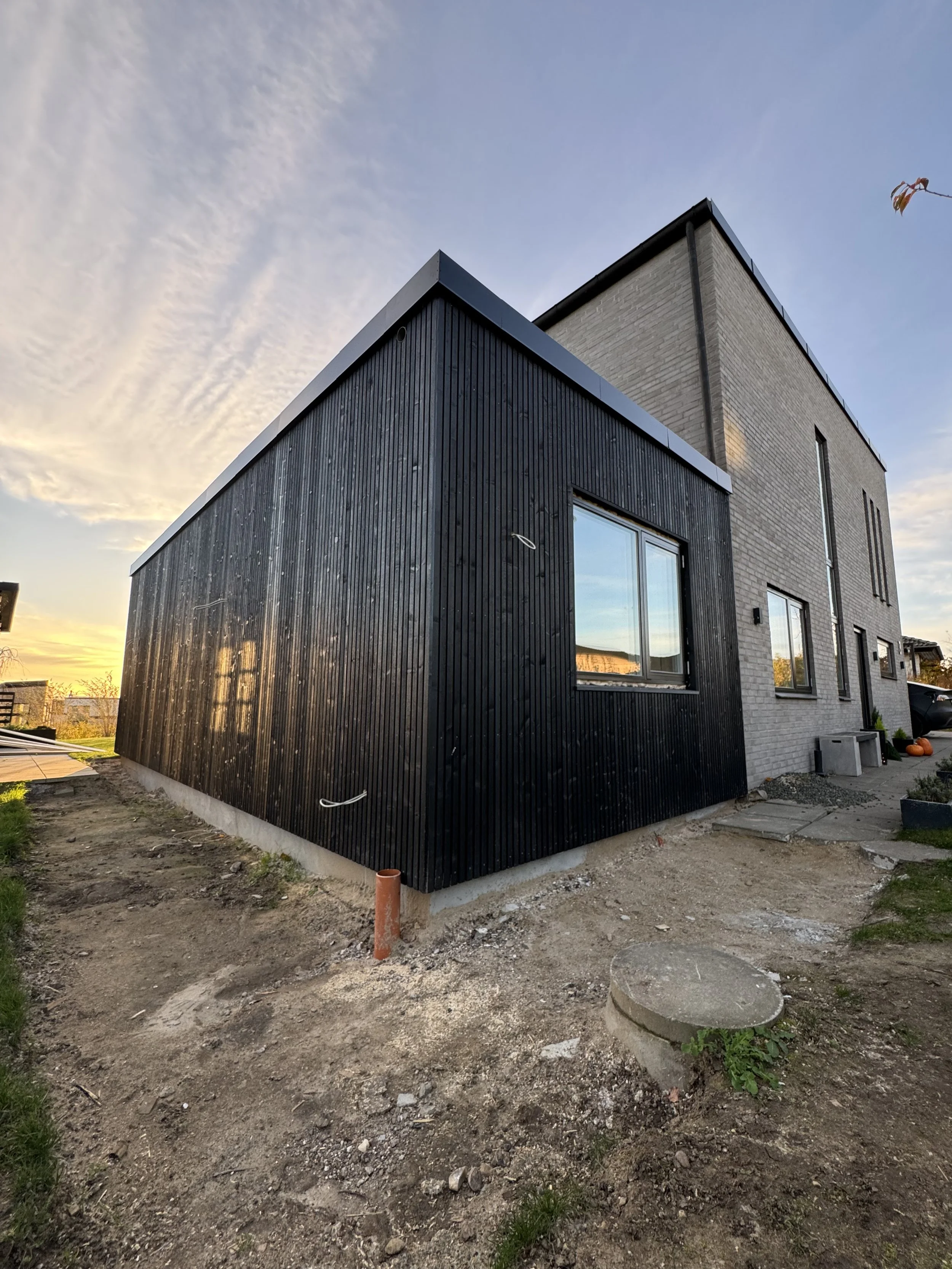 Et moderne byggeri med en sort træfacade på den ene side og en lys murstensfacade på den anden side, set udendørs ved solnedgang. Grunden er under anlæg, med jord og nogle byggeelementer omkring.