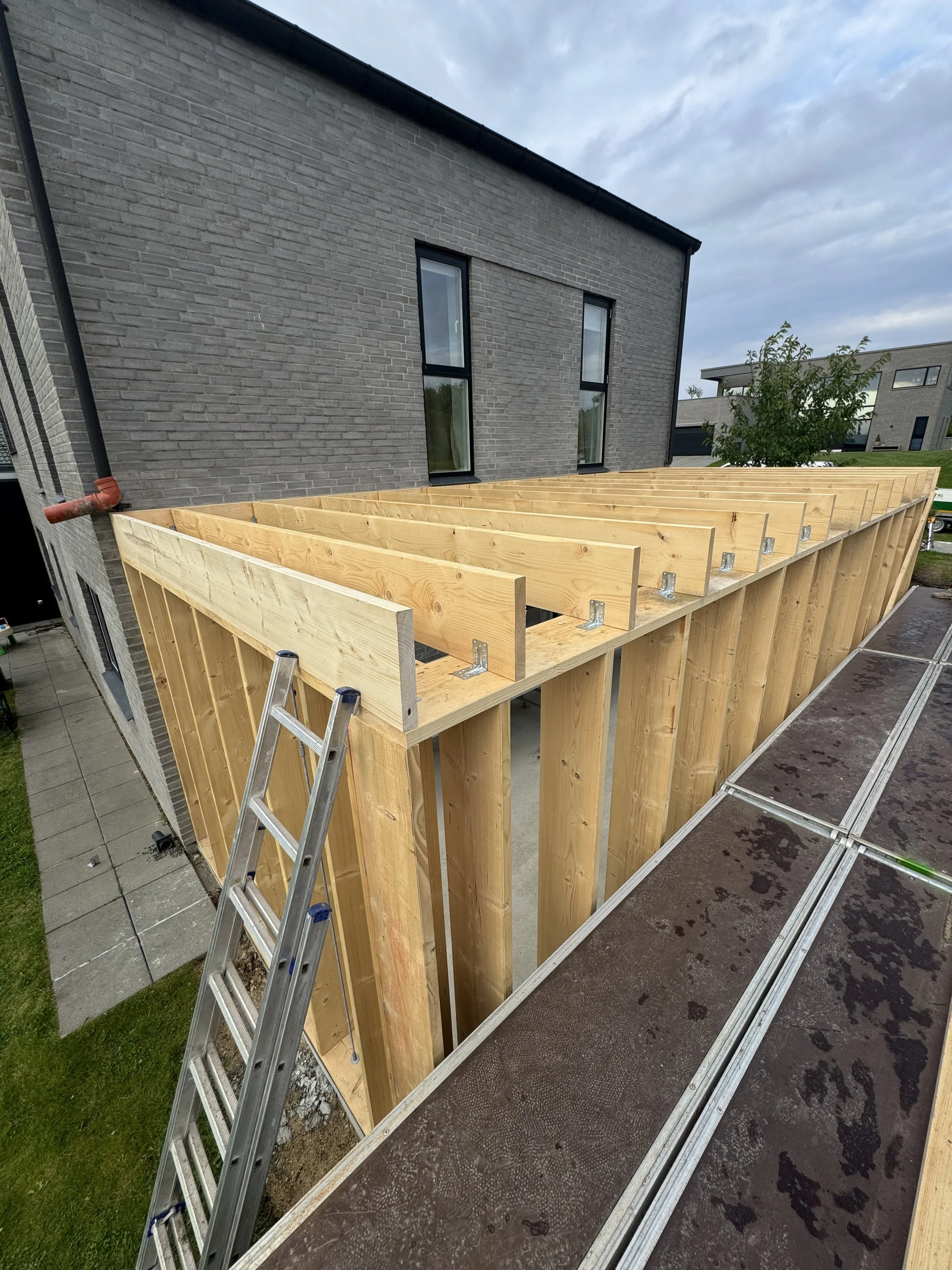 Byggeri af træværk på en terrasse foran en modern bolig med mørke vinduer og gråt murværk, med en stige og stillads i forgrunden.