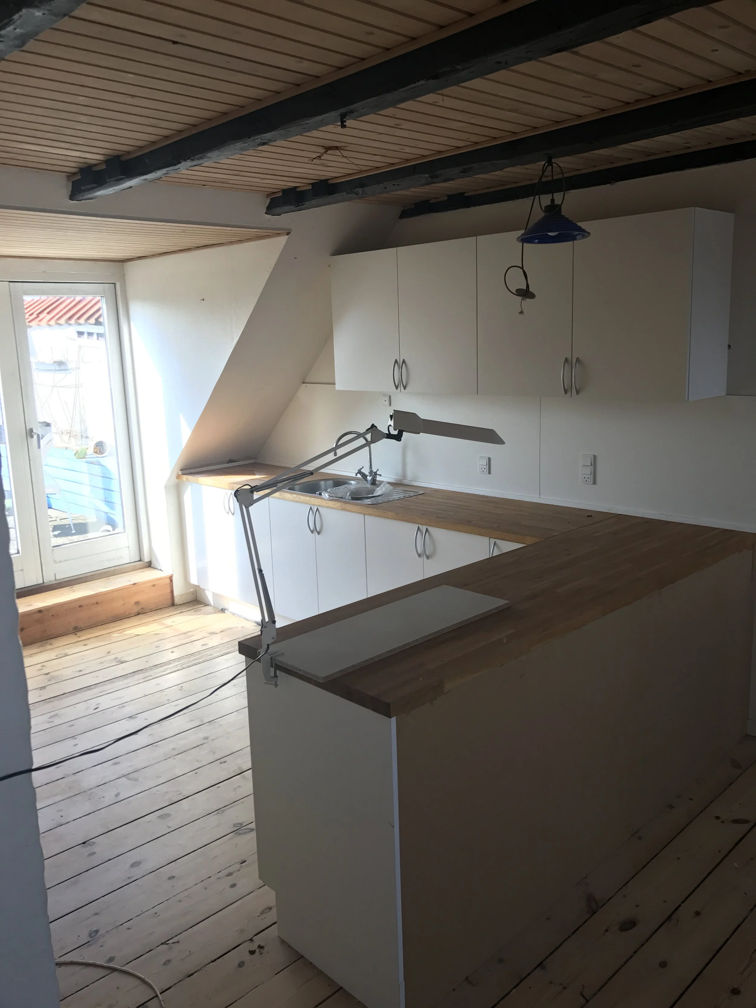 Køkken med hvide skabe, træbordplade, underopbevaringsskabe, og en arbejdslampe. Der er et stort vindue, der vender ud mod en terrasse, og trægulv.