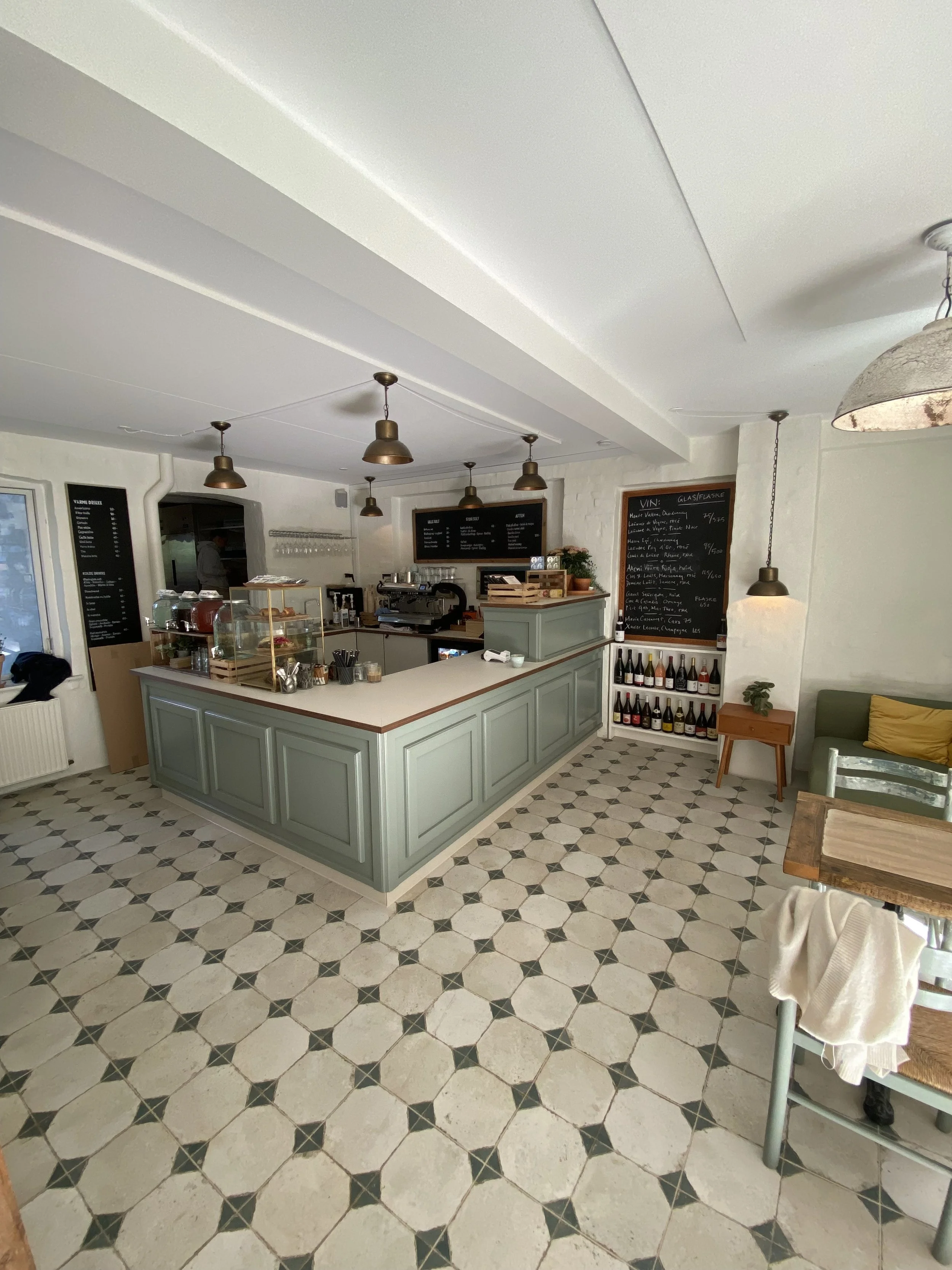 Indretning af en café eller restaurant med en disk i mintgrøn farve, sorte sorte tavler med menuer, og en sort og hvidmønstret terracotta gulv.