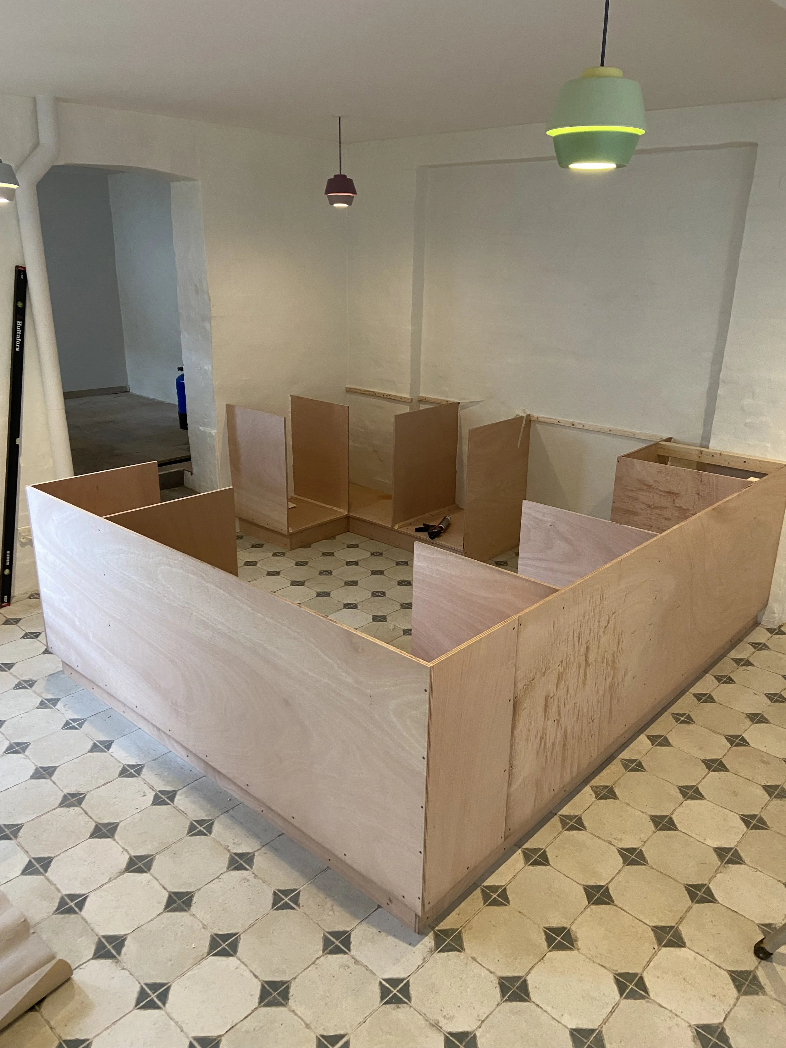 Et rum under renovering, med en lav konstruktion i træ, der sandsynligvis er en bar, og flere farvede lamper hænger fra loftet. Der er også værktøj og byggematerialer på gulvet.
