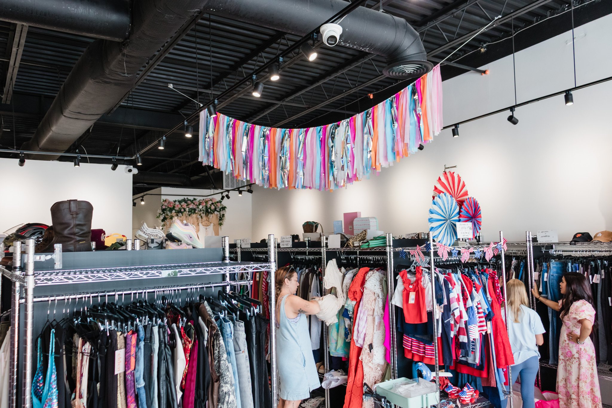 People shopping at a vintage, consignement/thrift store with clothing racks, colorful hanging decorations, and a boutique vibe