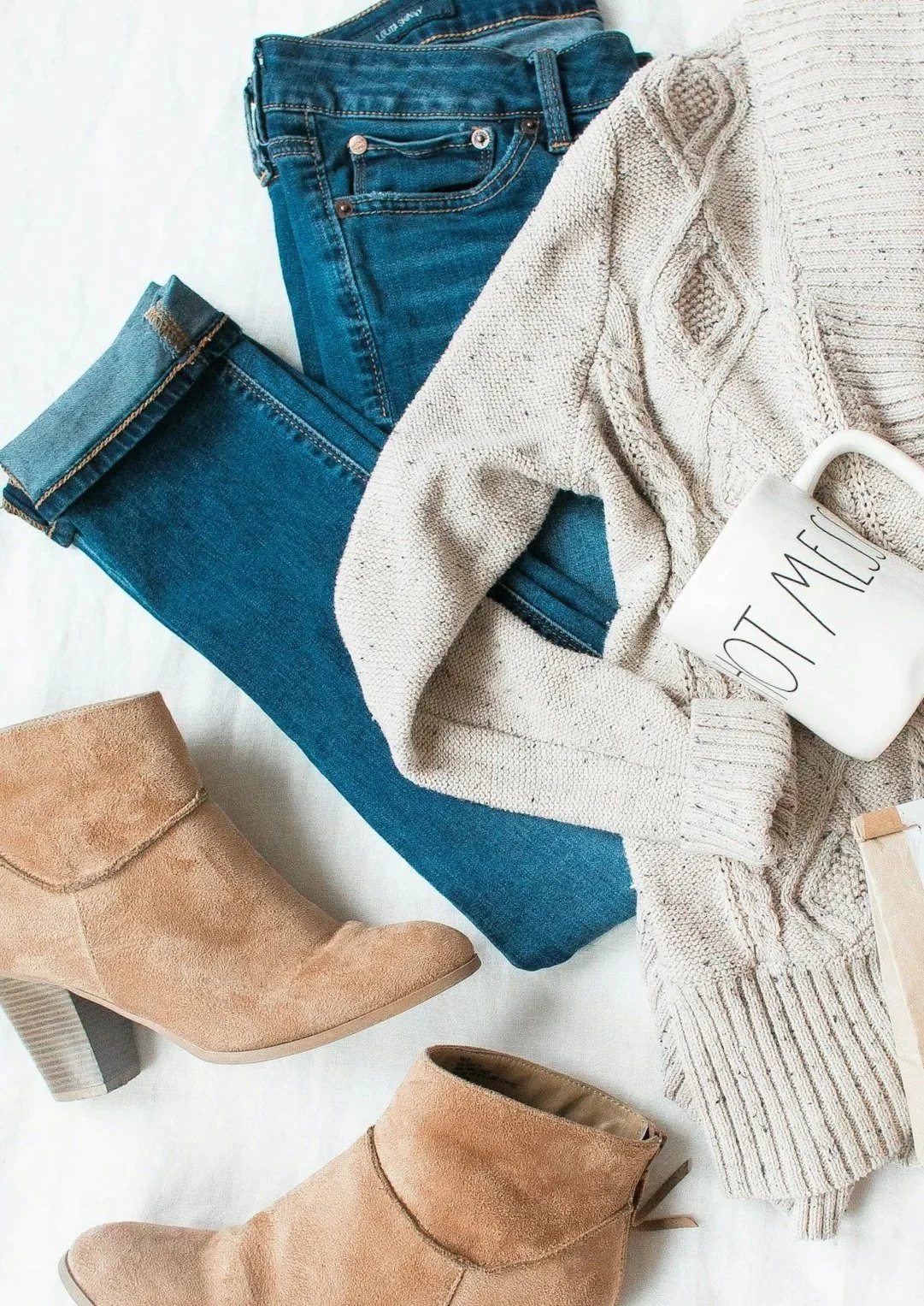 Pair of blue jeans, a cream-colored knitted sweater, tan ankle boots, and a white mug with black text.