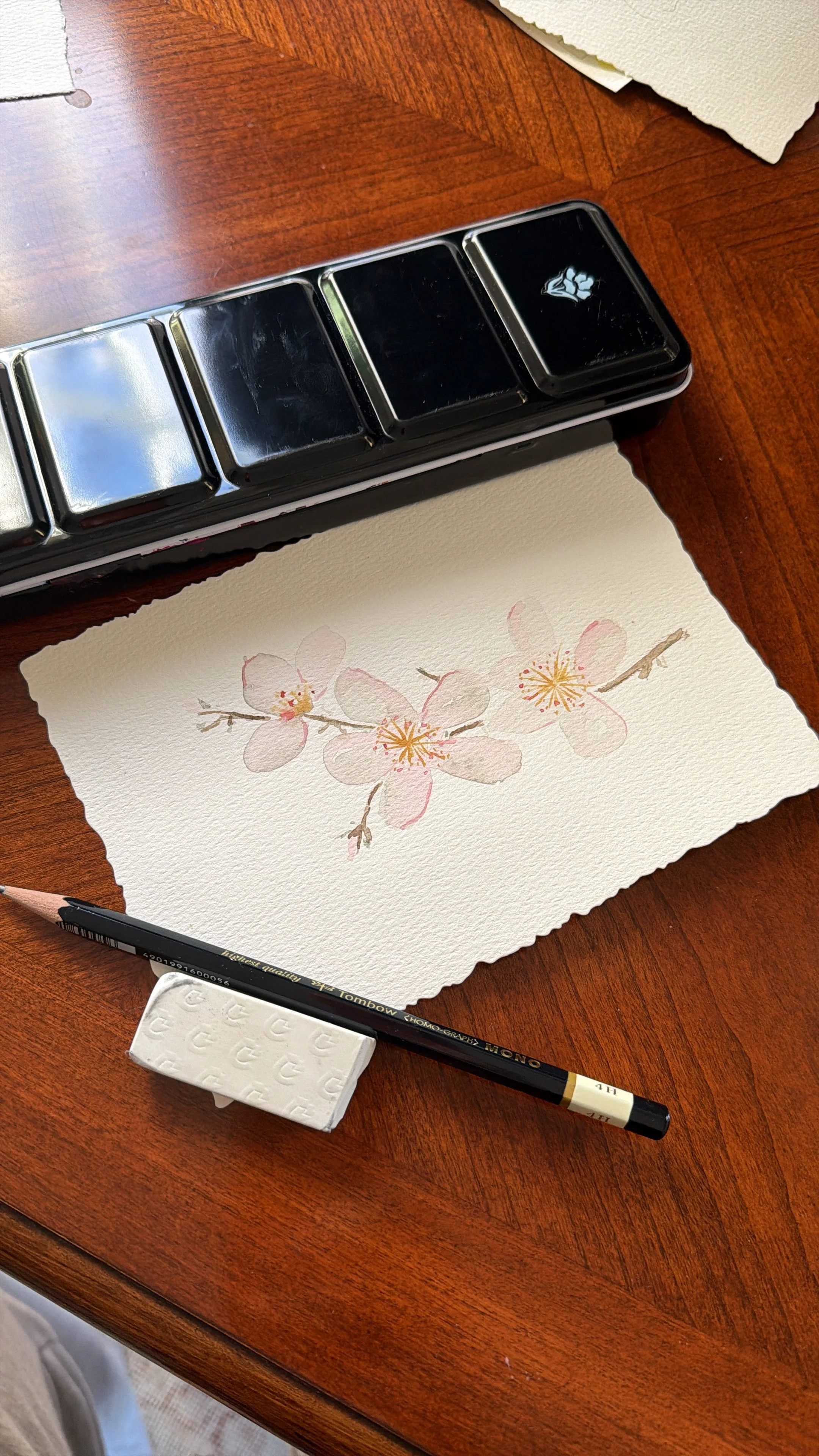 A watercolor painting of pink cherry blossoms on a branch, placed on a wooden table with a pencil, eraser, and ink pad nearby.