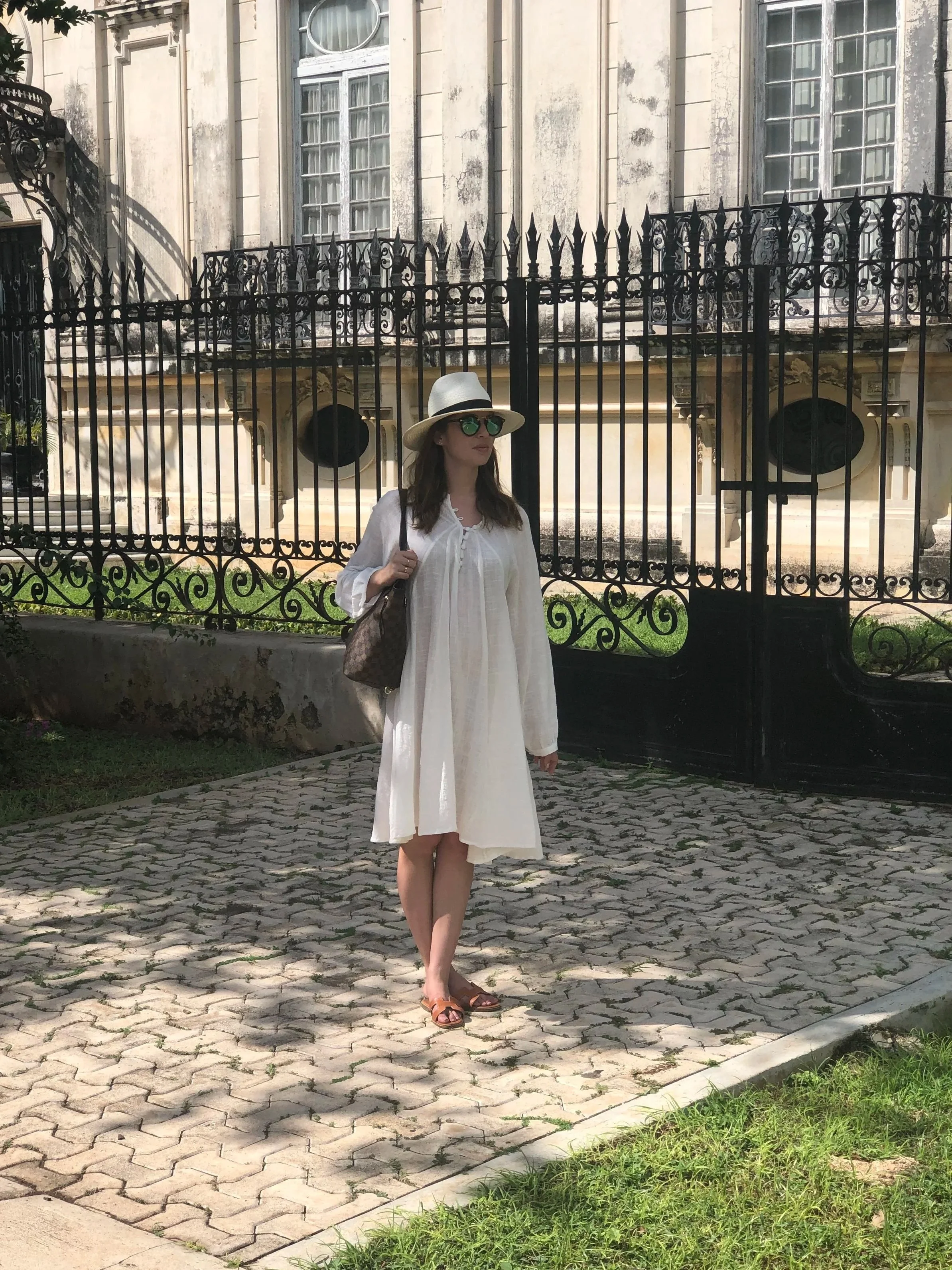A woman dressed in a white dress and sandals, wearing a white hat and sunglasses, standing on a paved walkway next to a black wrought iron gate and a historic building with tall windows.