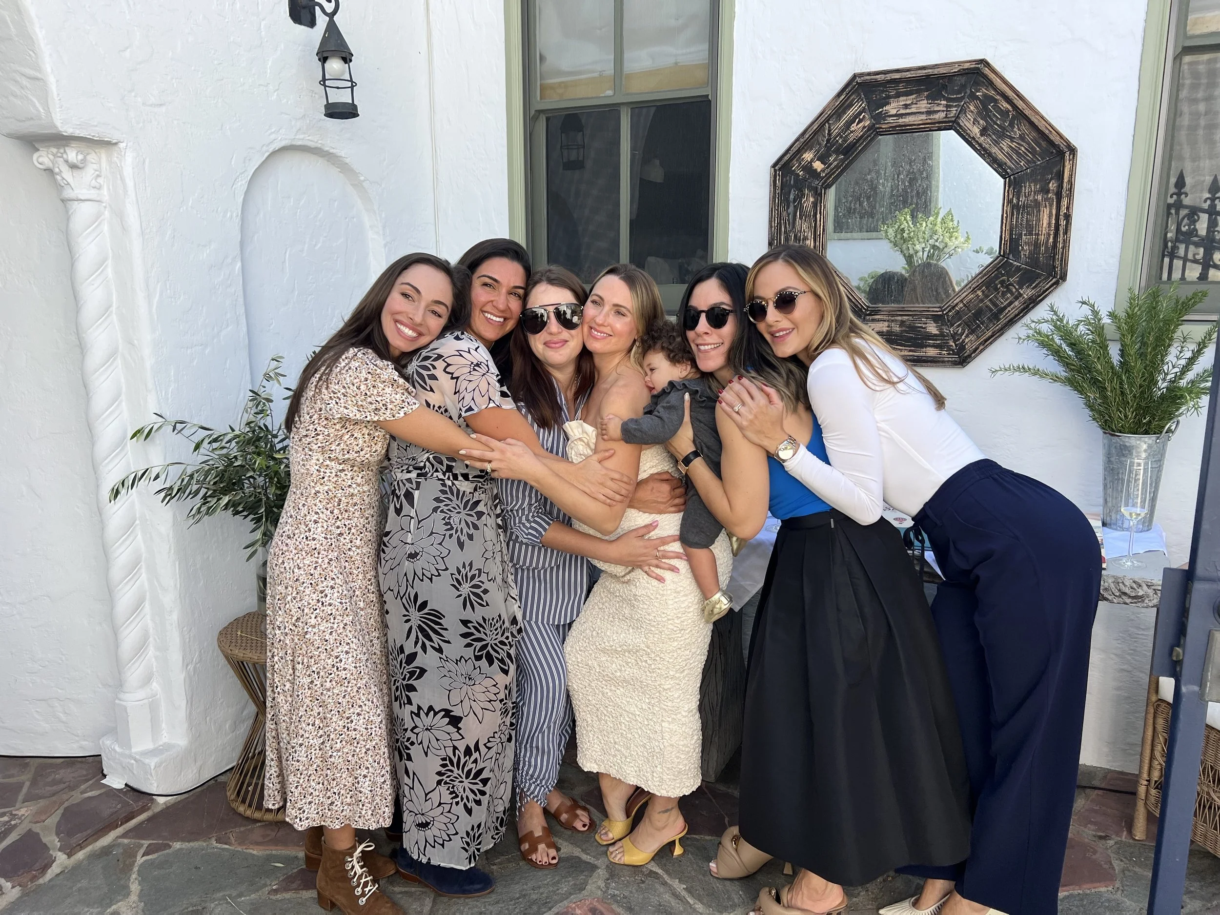 The boxing babes hugging and smiling outdoors in front of a white wall, with plants and a mirror in the background at a baby shower.