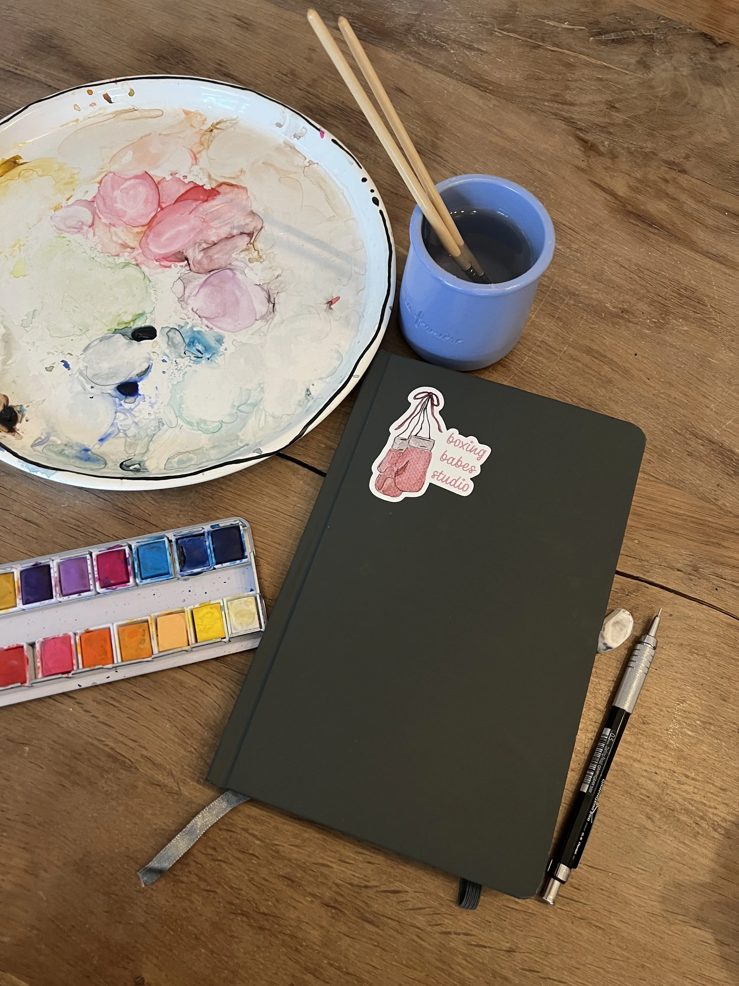 A wooden table with a watercolor palette, a notebook with a boxing-themed sticker, a pen, a small cup of black ink with chopsticks inside, and a paintbrush cap.