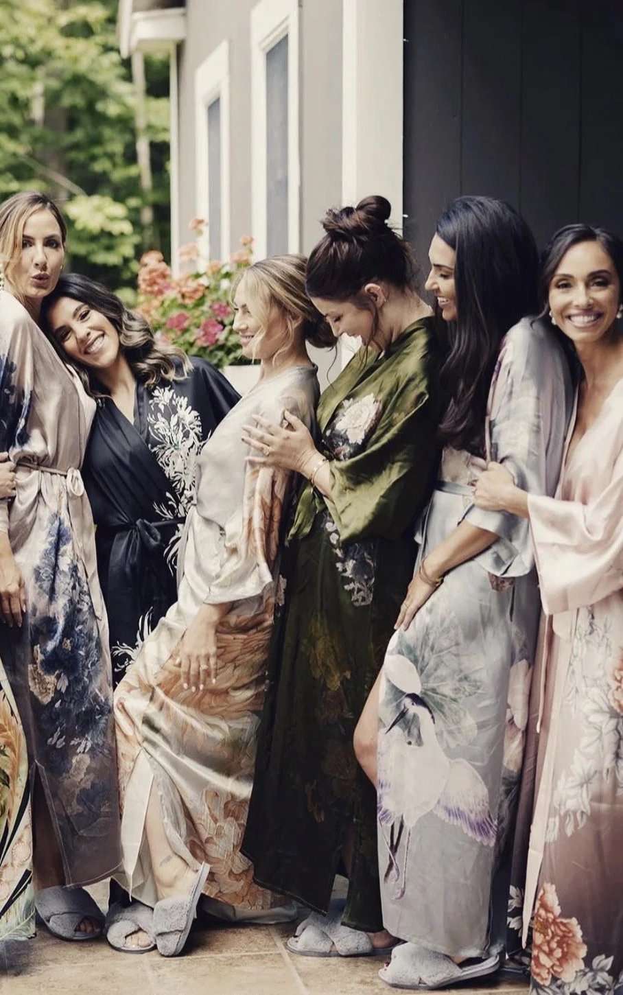 The boxing babes wearing silk robes with floral and bird patterns, standing together outside on a patio with greenery and flowers in the background getting ready for a wedding.