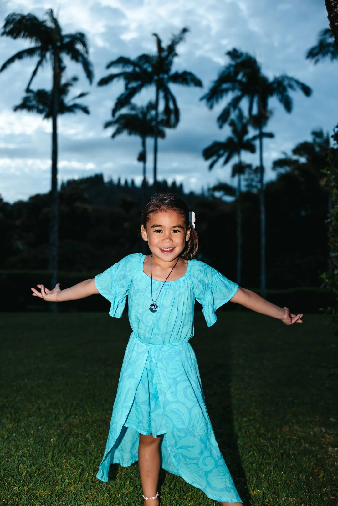 A young girl in a blue dress standing outside on grass with palm trees and a cloudy sky in the background, smiling with arms outstretched.