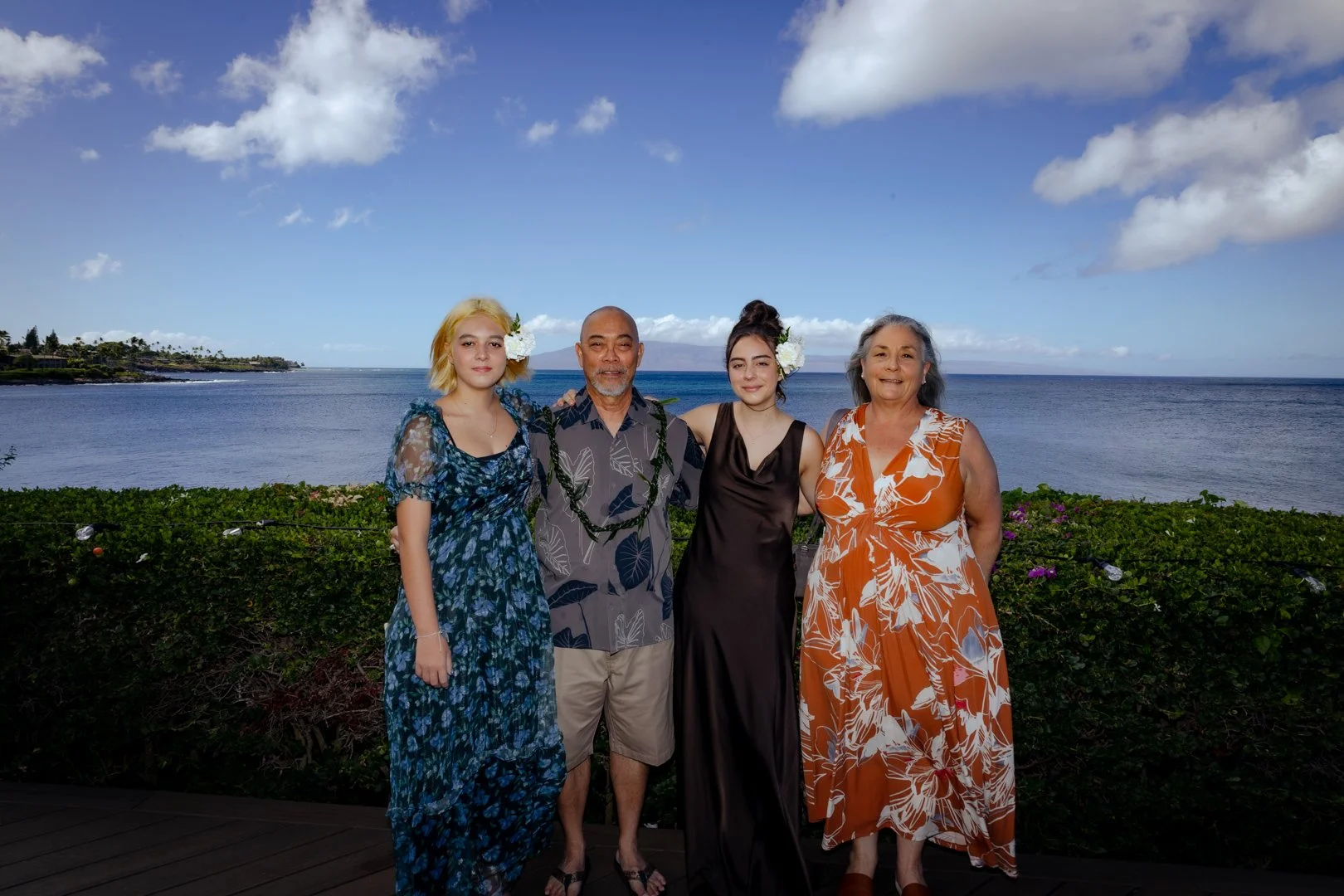 Four people standing together near the ocean on a clear day, with two women wearing floral dresses and two men in casual shirts and shorts, behind green shrubbery and ocean water.
