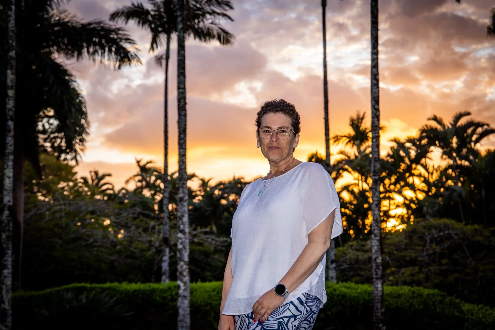 A woman standing outdoors at sunset, with palm trees and lush greenery in the background.