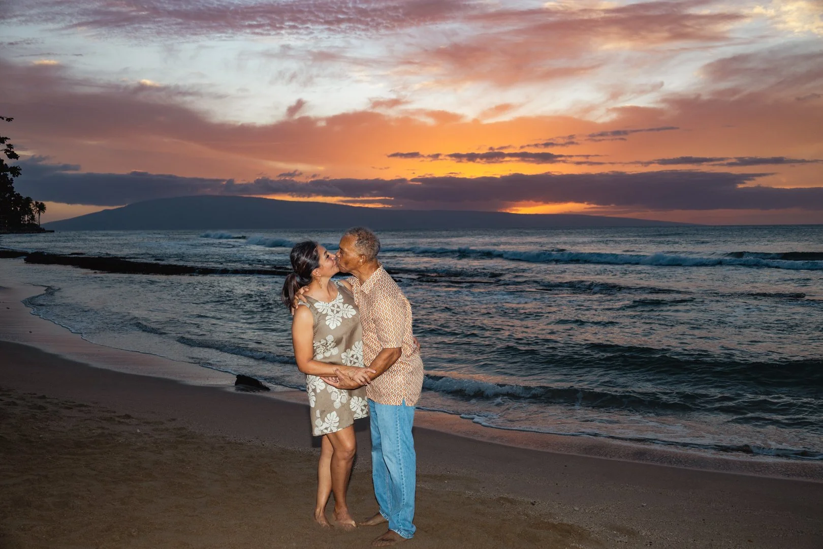 An elderly man and a woman kissing on a beach at sunset, with water and clouds in the background.