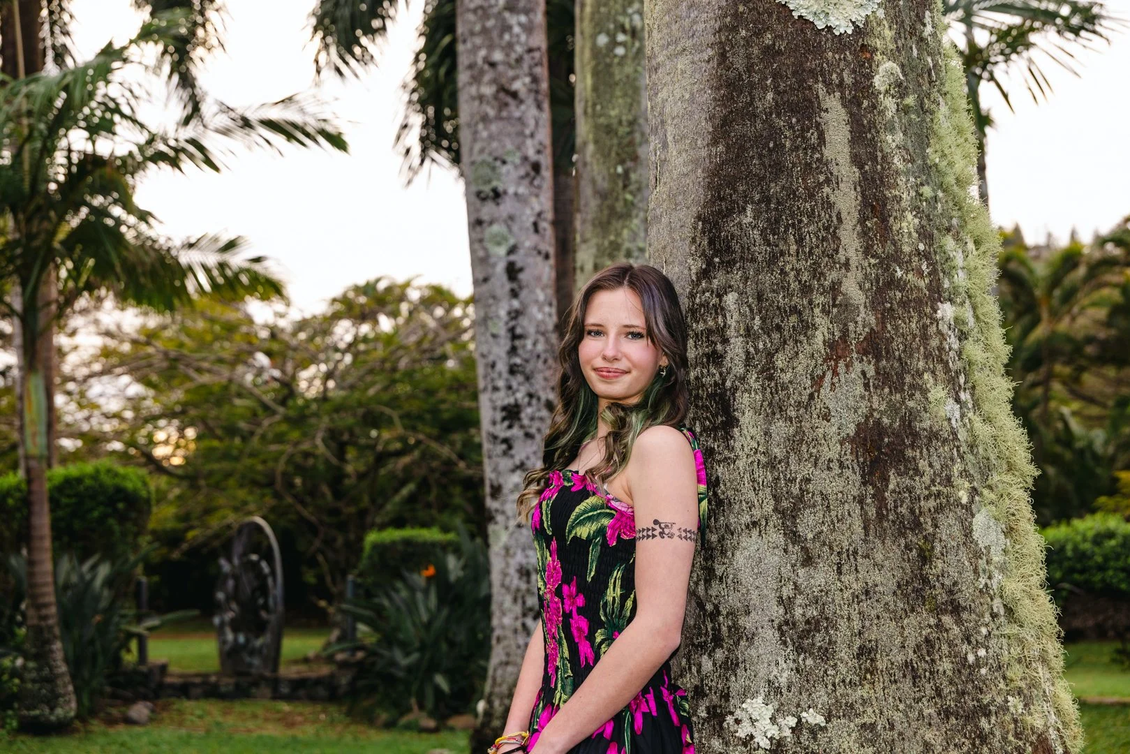 A young woman with long brown hair and green highlights, wearing a sleeveless black dress with pink floral patterns, stands next to a large tree trunk in a lush garden with green trees and plants in the background.