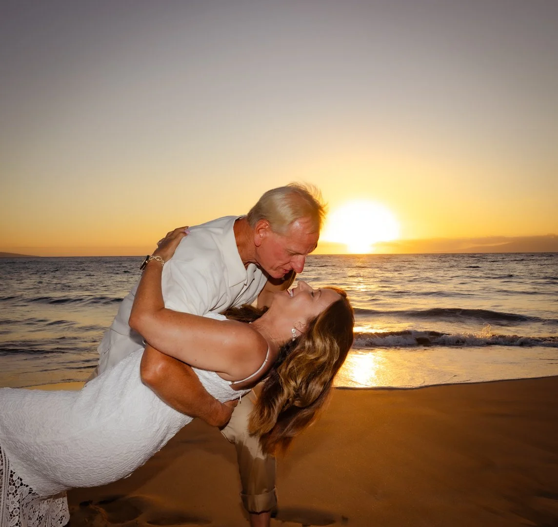 A senior man and a woman are dancing on a beach at sunset, with the man dipping the woman as they smile at each other.