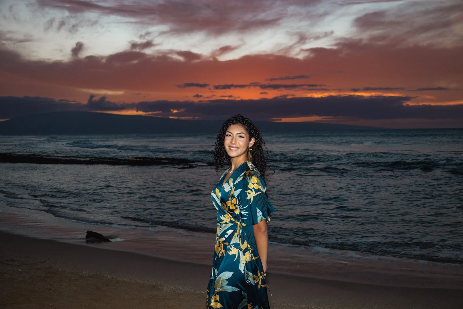 A woman in a floral dress standing on a beach at sunset, smiling with the ocean and sky in the background.