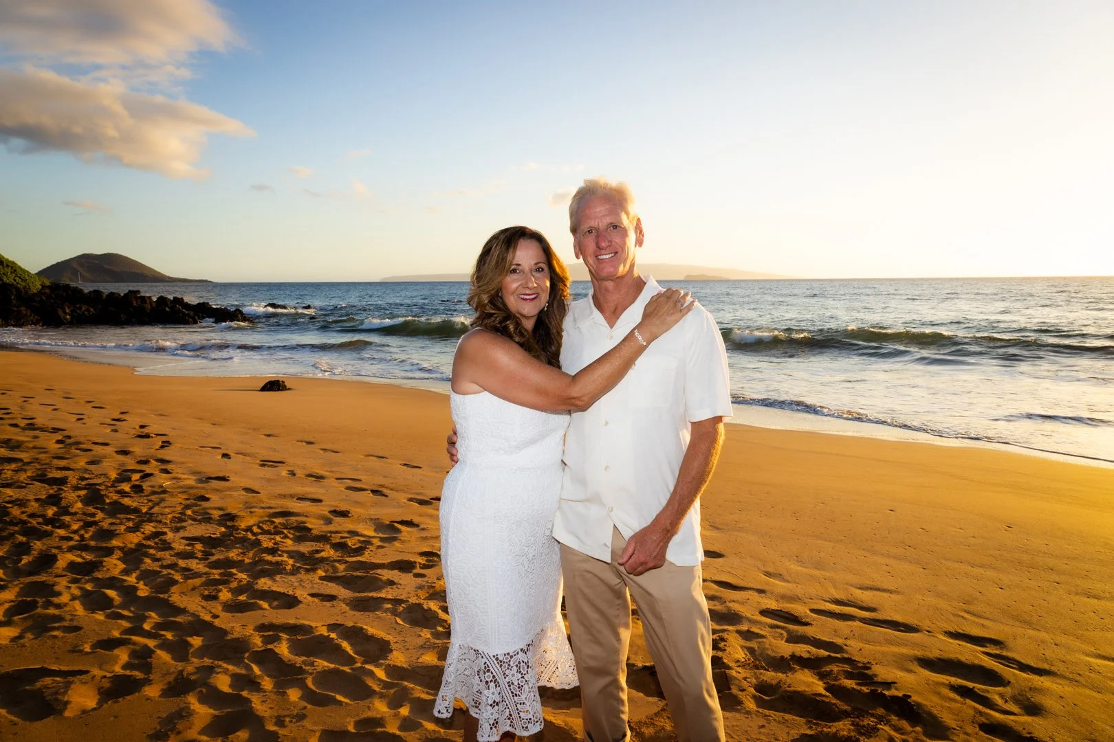 A smiling couple stands on a sandy beach with the ocean and distant hills in the background during sunset, embracing each other.