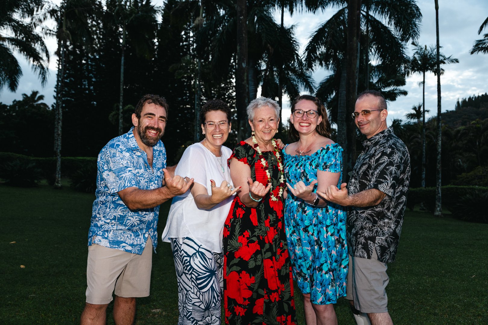 Group of five people outdoors dressed in Hawaiian-style clothing, smiling, with trees in the background.